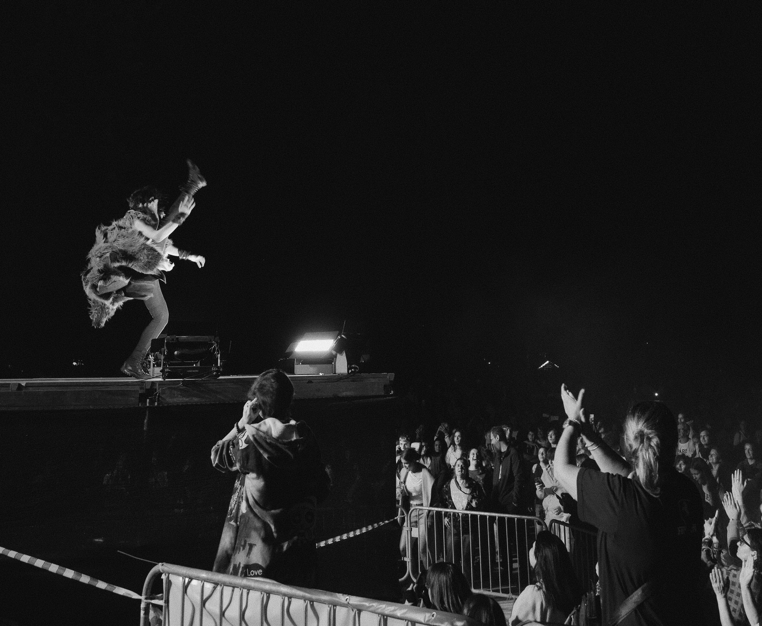 A performer on stage at night kicking a person with long hair into the air, with a crowd of fans cheering and taking photos behind a safety barrier. The scene is in black and white.