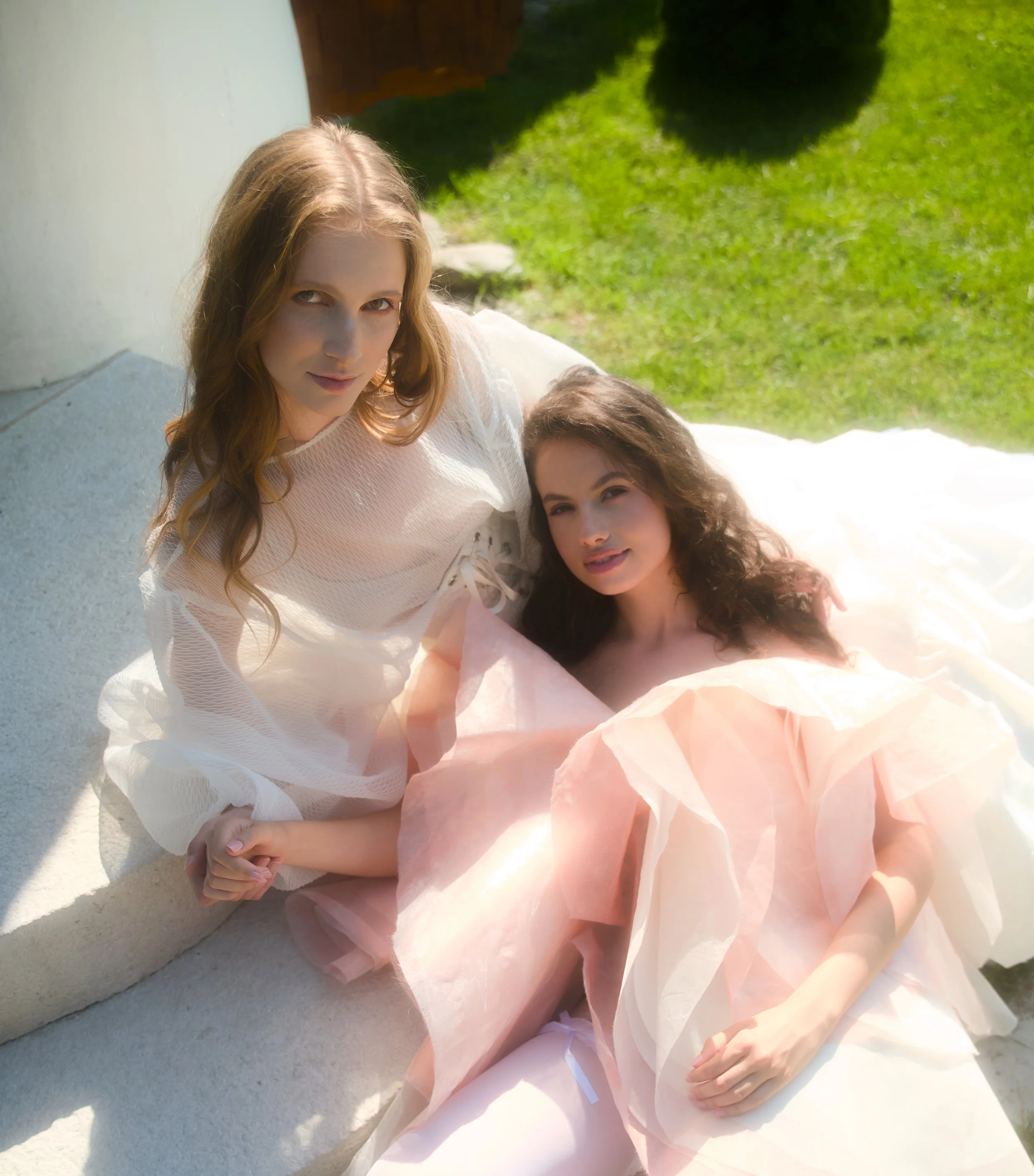 Two young women in pastel dresses sitting on a white surface outside on a sunny day, with a green lawn in the background.