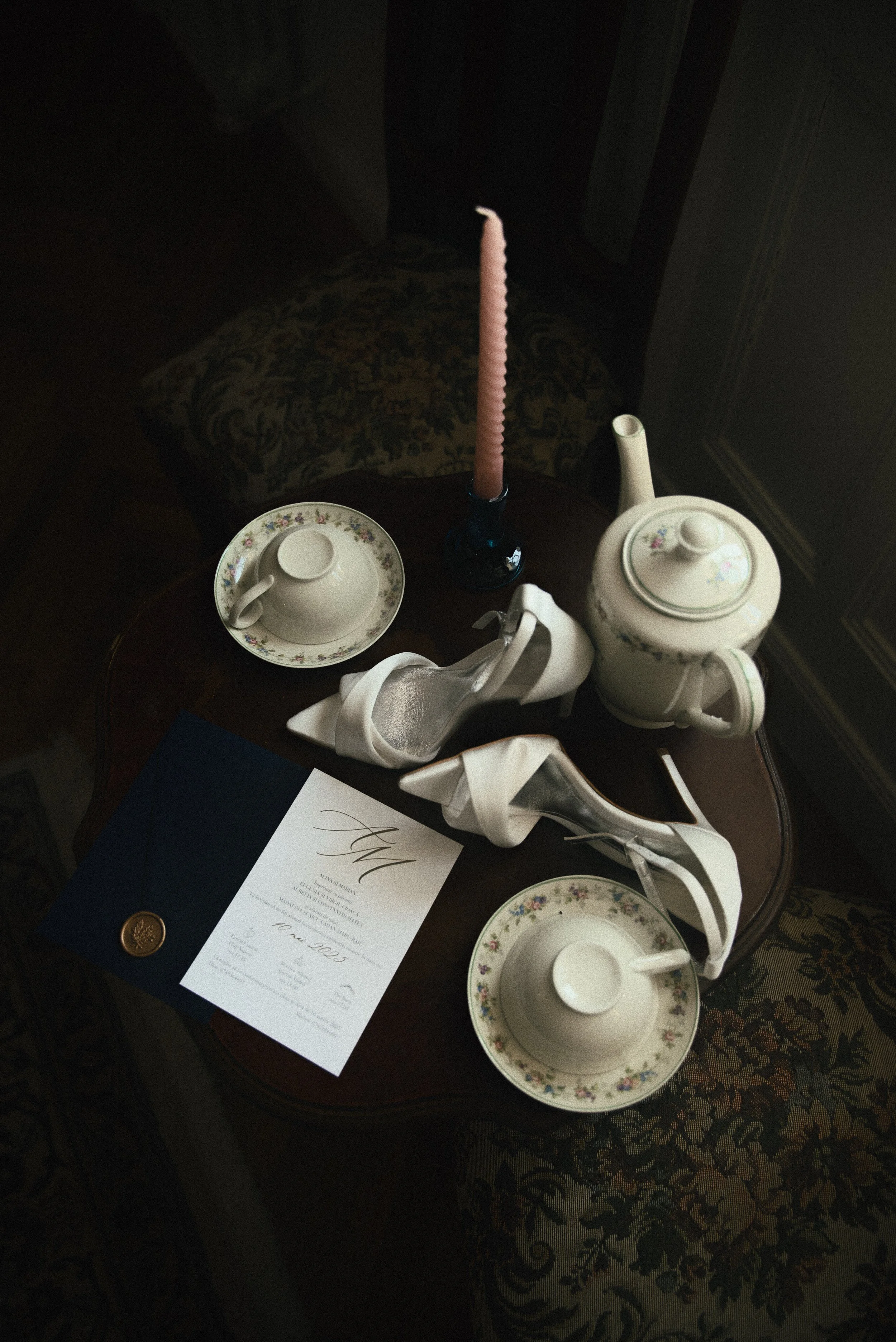 A vintage tea setting on a wooden table includes a teapot, cups and saucers, a pair of white high-heeled shoes, a pink taper candle in a blue holder, and a formal invitation letter with a gold seal.