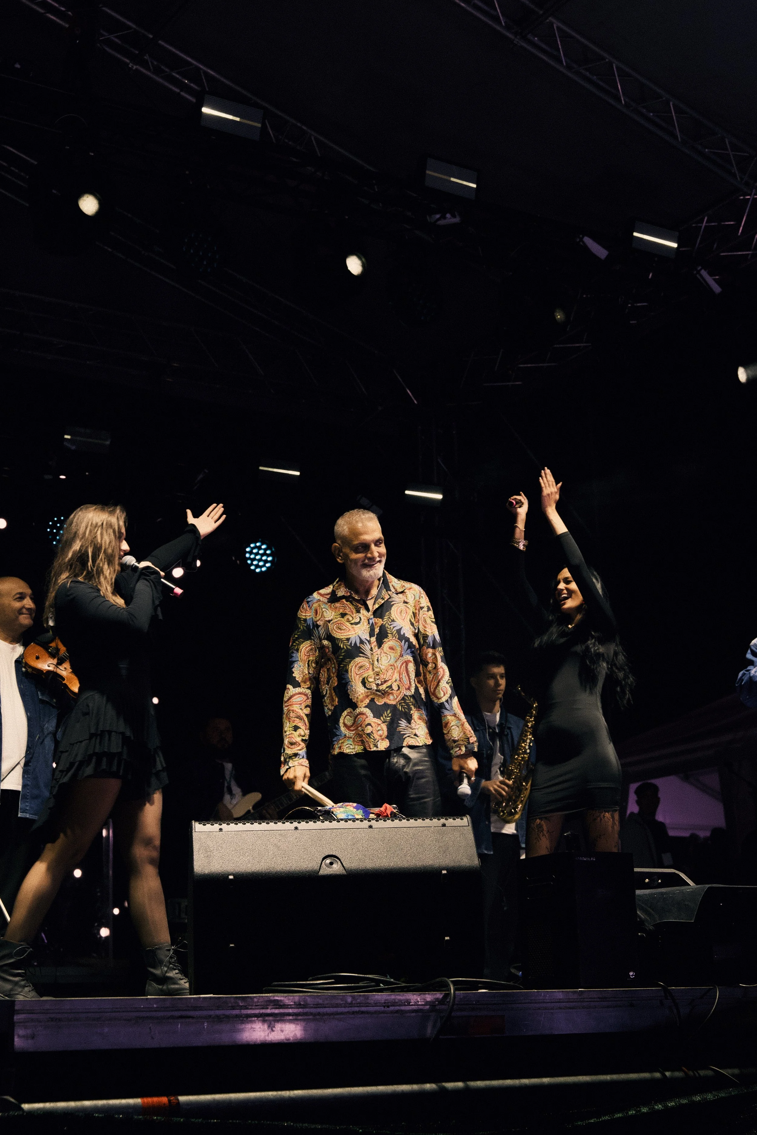 People on stage during a live music performance, with one woman singing into a microphone, a man holding a drumstick, and other musicians in the background, under stage lighting.