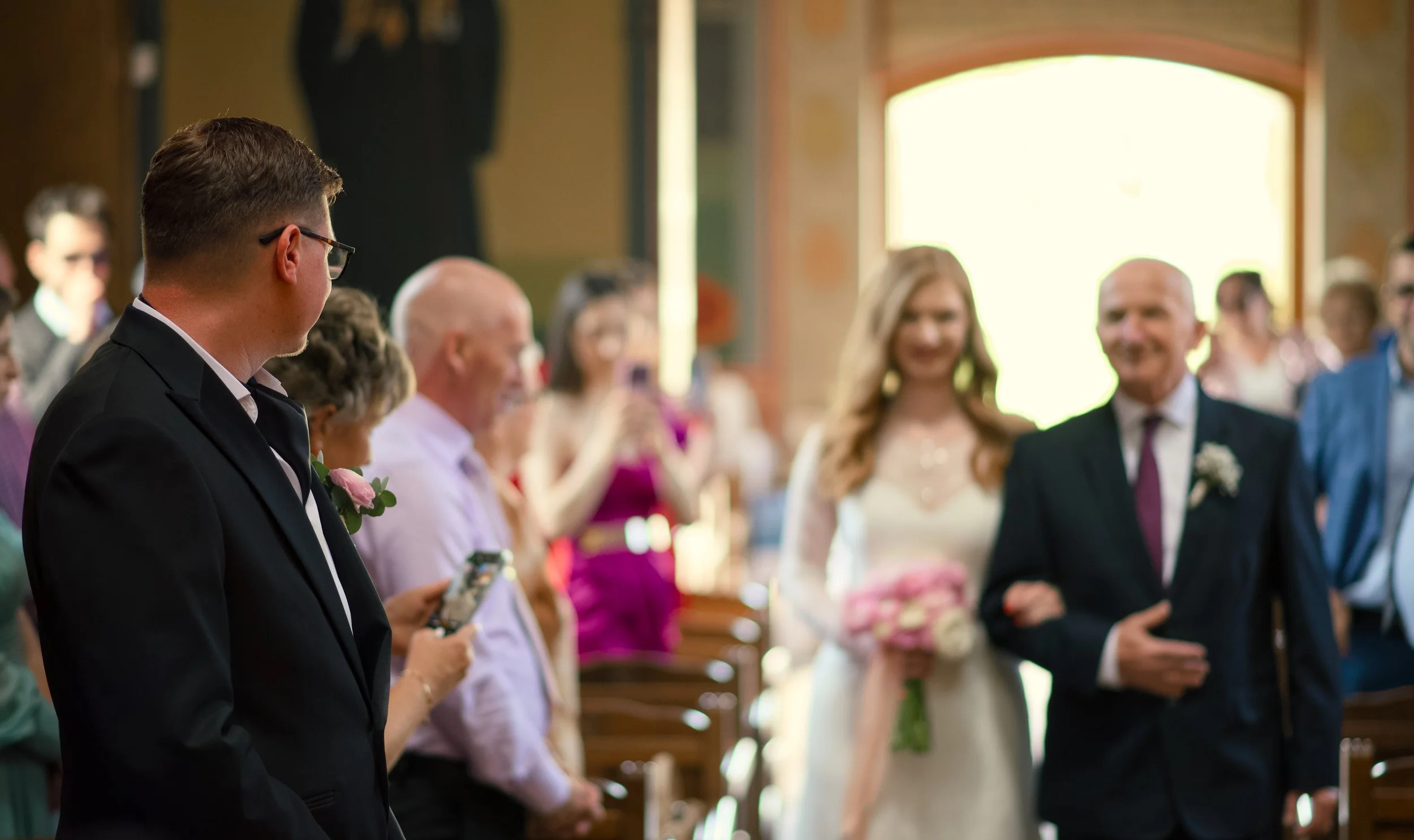A groom in a black suit looking at a bride walking down the aisle in a white dress holding a pink bouquet, as family and friends watch during a wedding ceremony in a church.