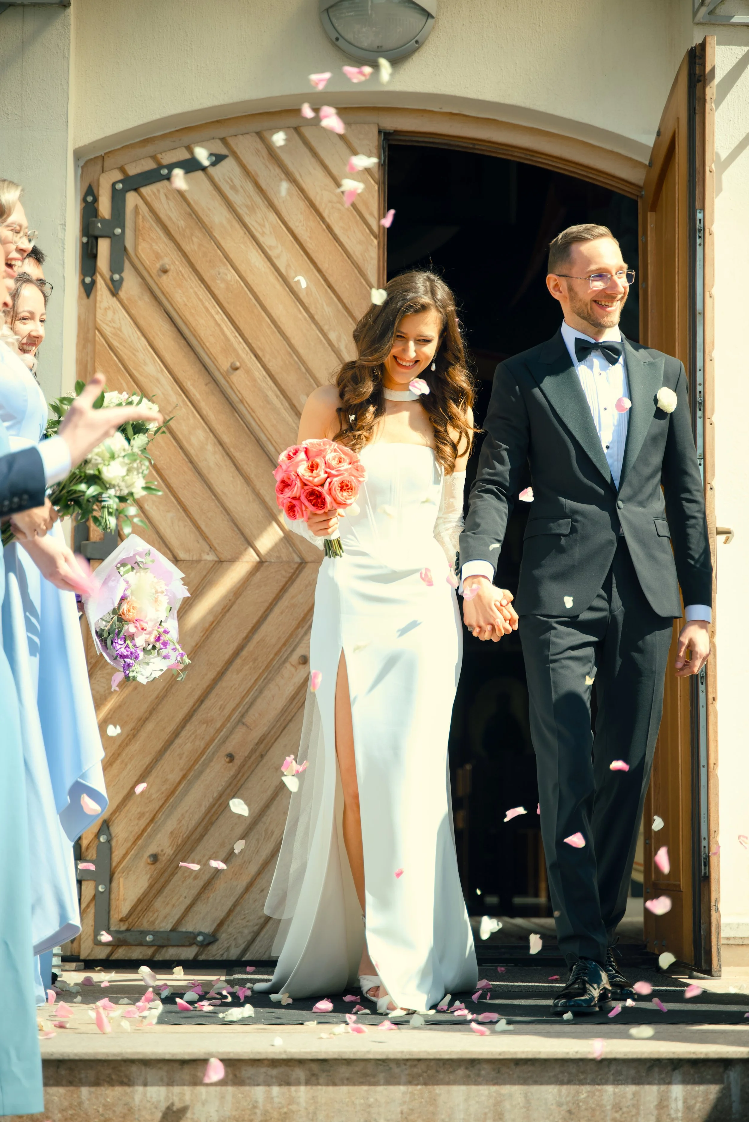 A newlywed couple, the bride holding a bouquet of pink and peach roses, holding hands with the groom, as they leave a church or ceremony venue. Bridesmaids and guests throw flower petals in celebration.