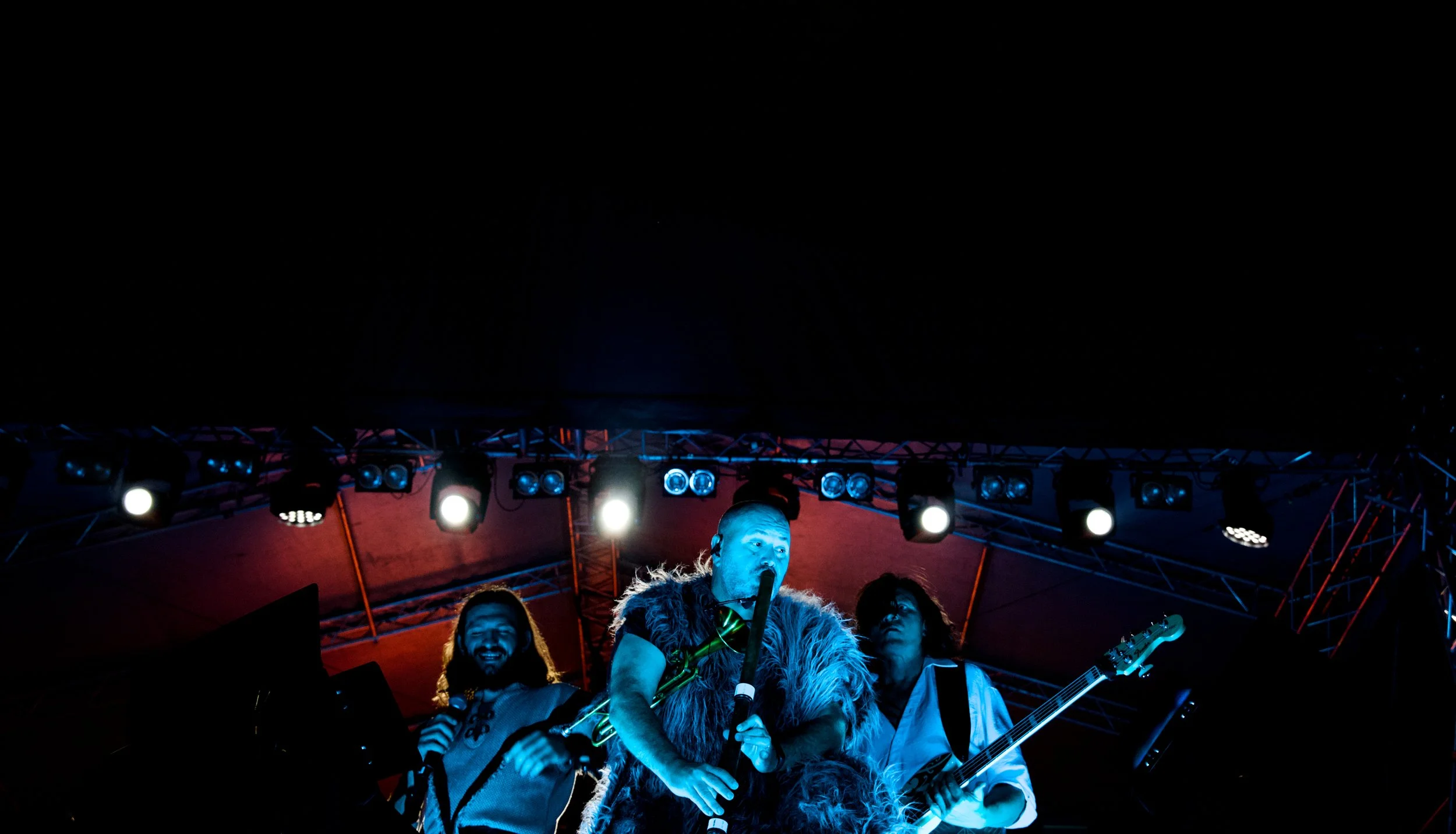 Band performing on stage with three musicians, one singing into a microphone and playing a wind instrument, another playing bass guitar, all illuminated by stage lights against a dark background.
