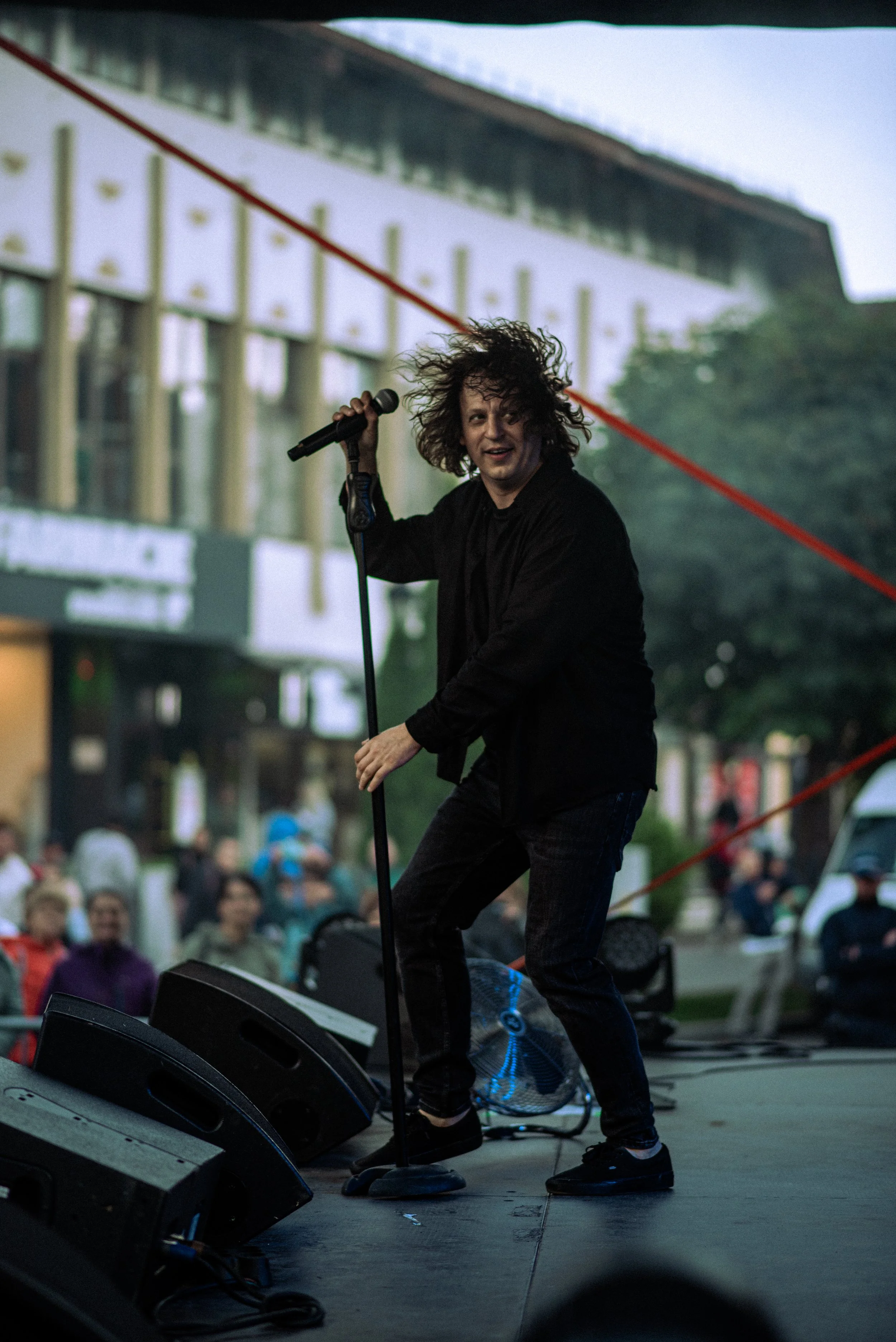 Performer with curly hair holding a microphone on stage during outdoor concert with audience and city building in background.