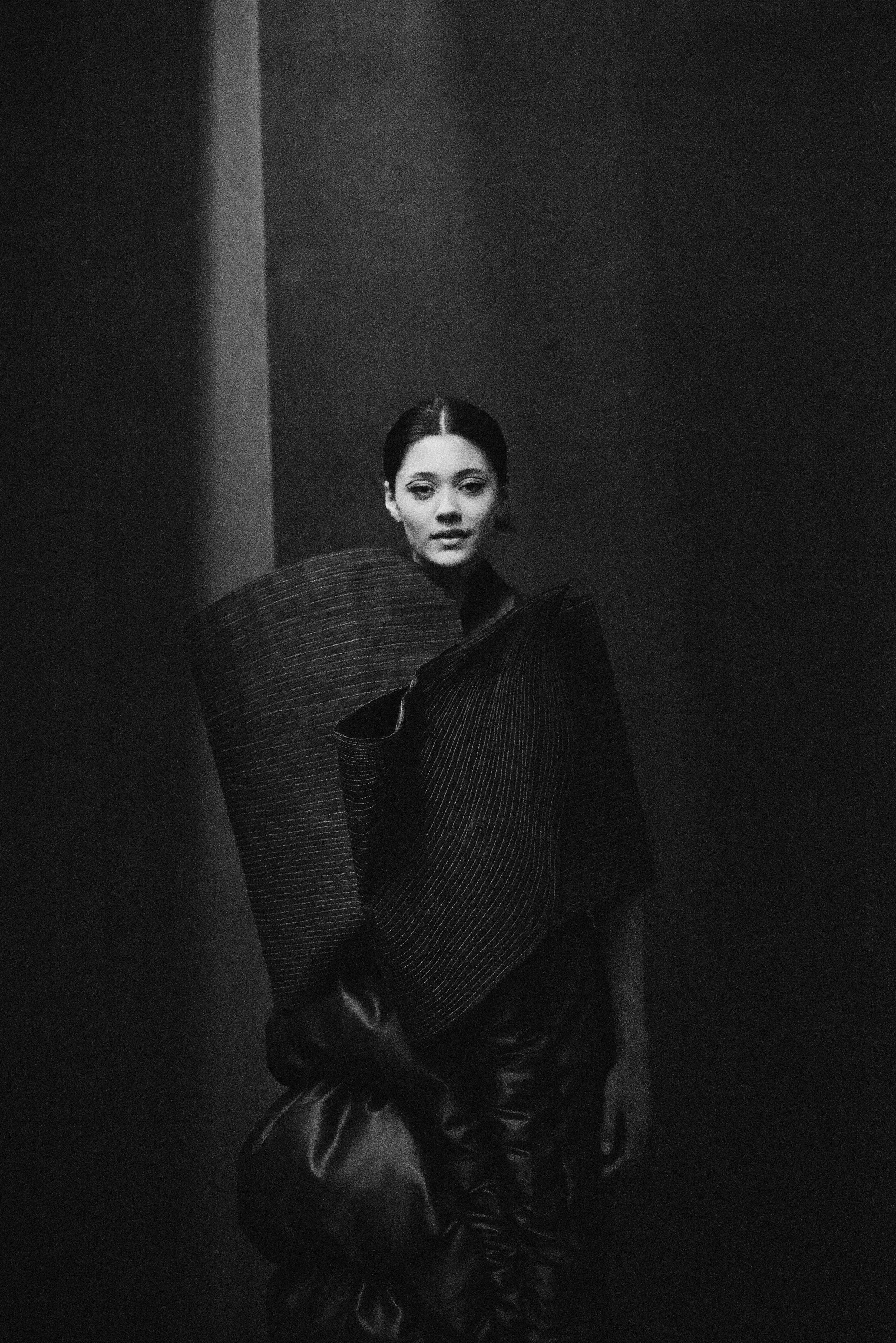 A black and white photograph of a woman with short dark hair, wearing an avant-garde black outfit with large, textured, sculptural elements on her shoulders and arms, standing against a dark background.