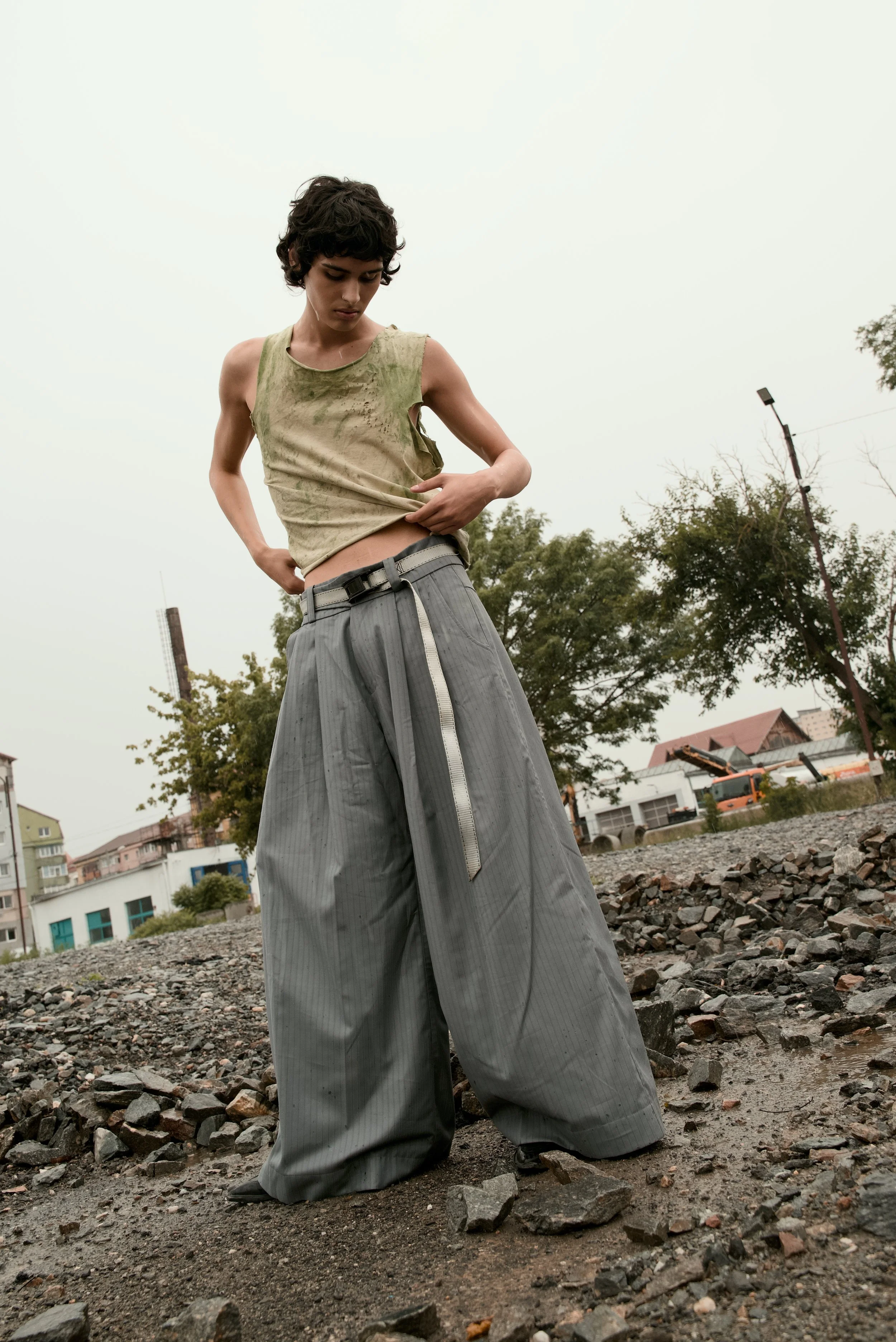 Young person with short curly hair standing outdoors on rocky ground, wearing a dirty sleeveless top and loose oversized gray pants.