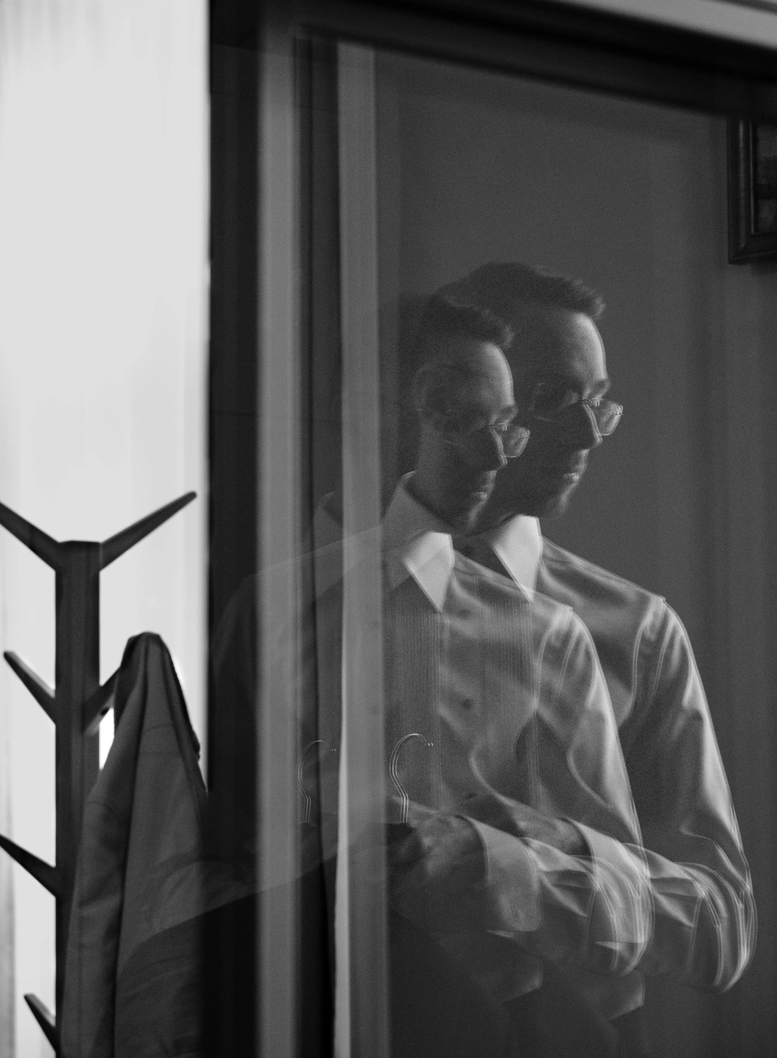 A black and white photo of a man with glasses, wearing a button-up shirt, looking at his reflection in a mirror. The reflection shows him with arms crossed. A coat rack with a jacket is visible in the foreground, and the room has a plain wall with a