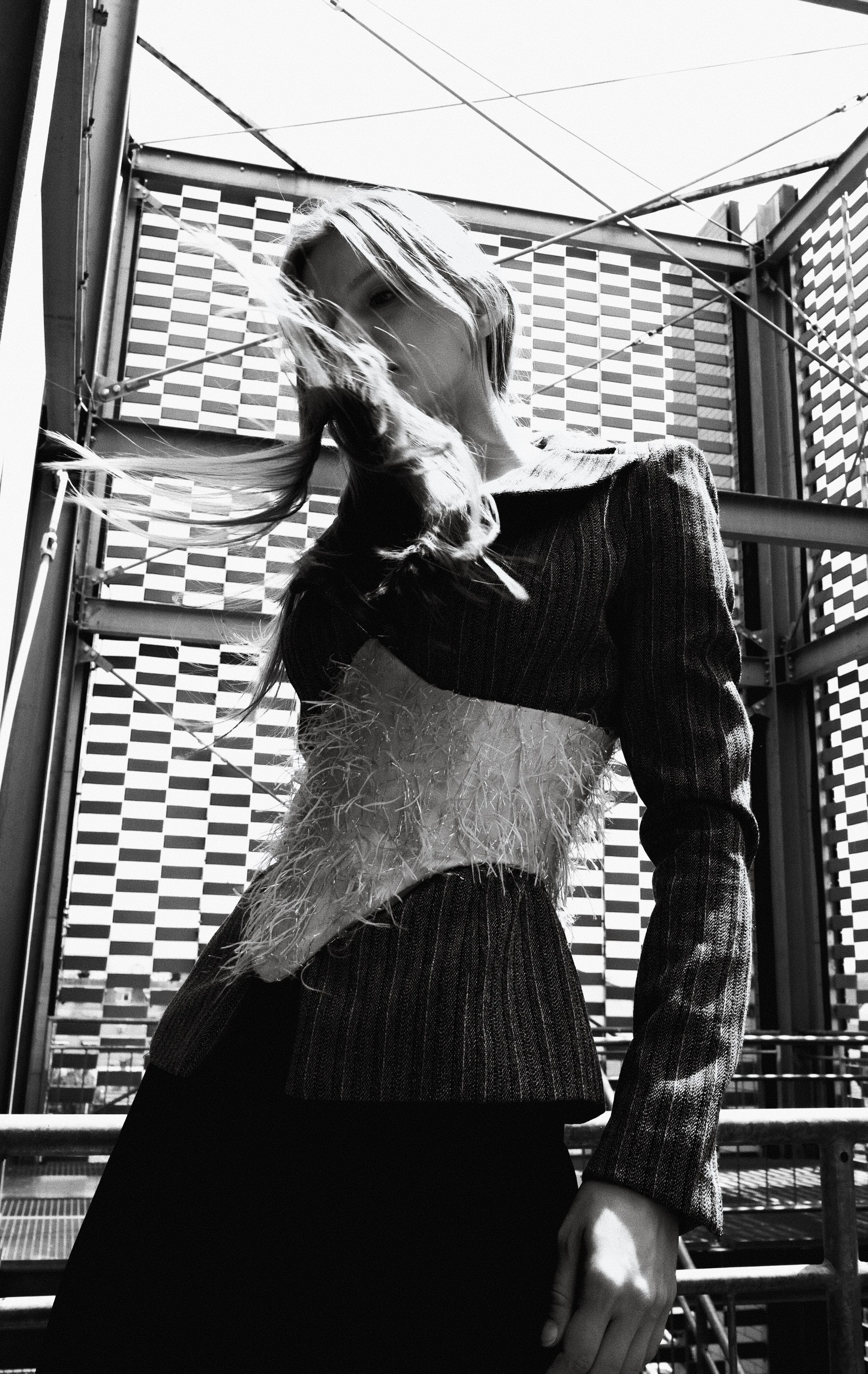 A black and white photo of a woman with long hair in wind, wearing a textured top with feather details, standing in an outdoor urban setting with a building and sky in the background.