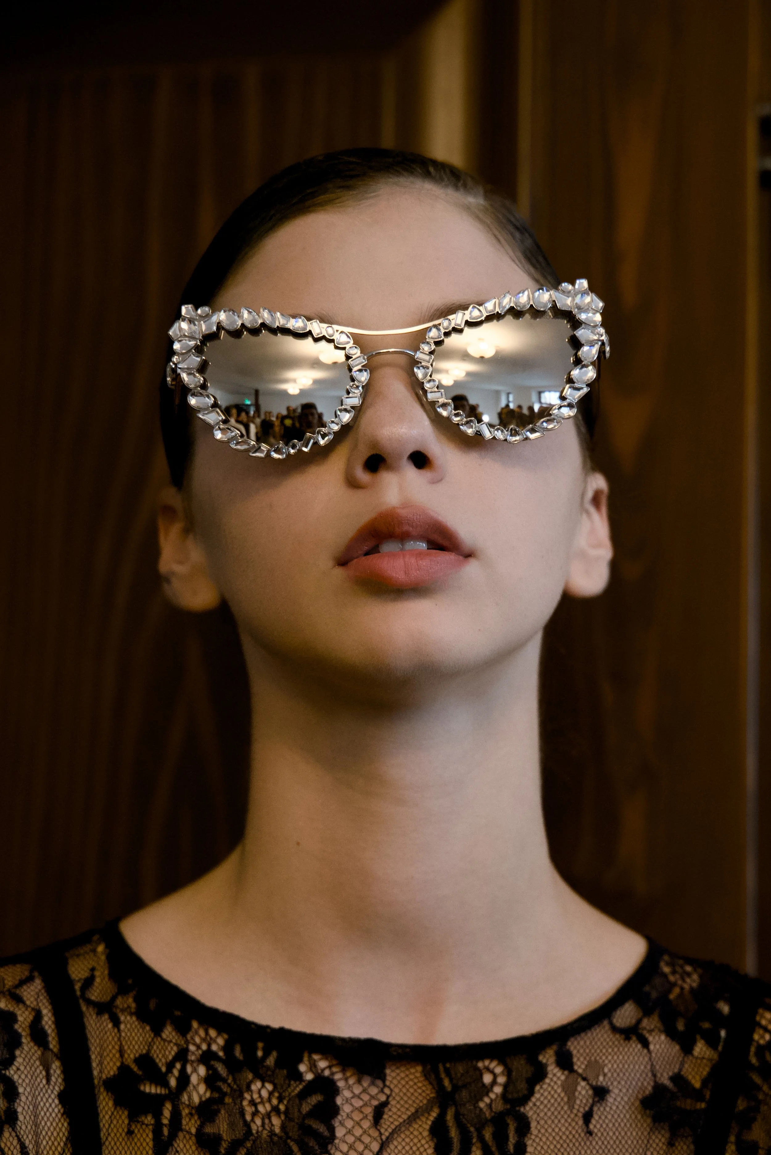 Close-up of a woman with fair skin and dark hair wearing large, reflective sunglasses with a decorative rim, and a black lace top.