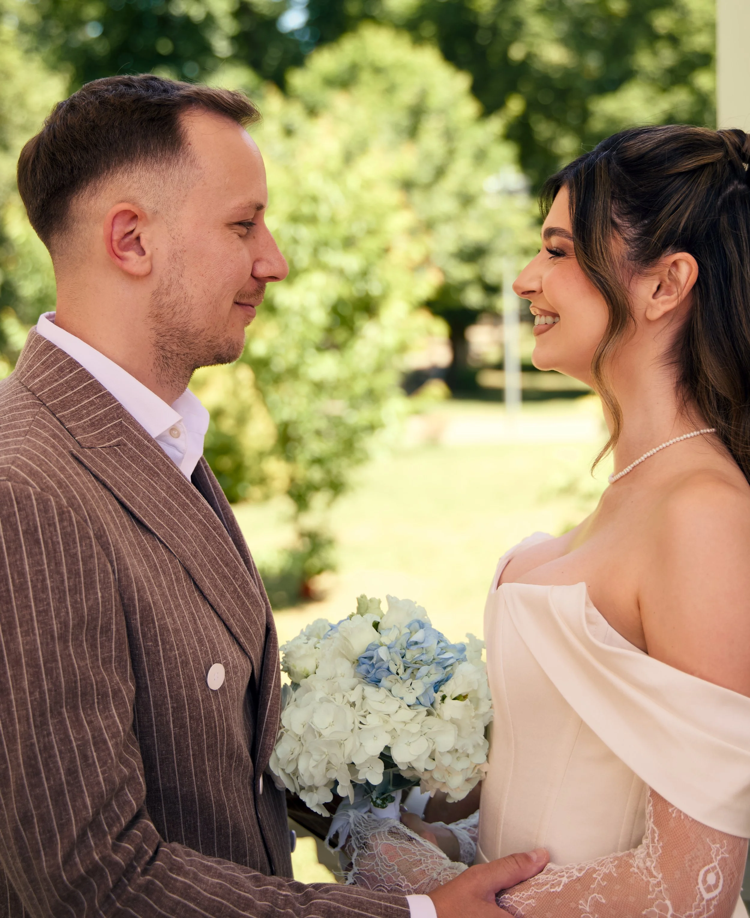 A newlywed couple stands face to face outdoors, smiling, with the groom holding a bouquet of white and blue flowers.