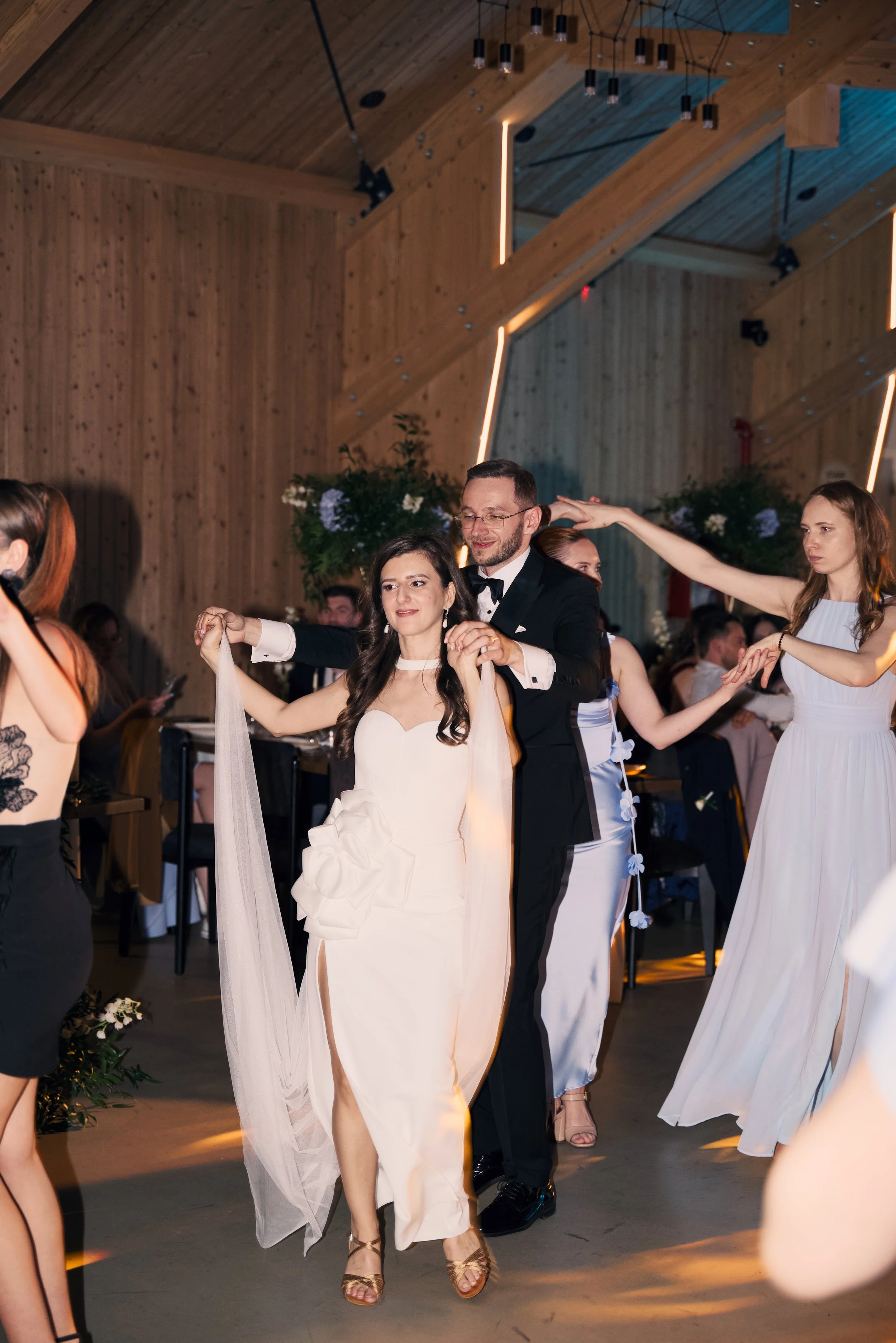 A wedding reception with a bride and groom dancing together surrounded by bridesmaids and guests in a decorated wooden hall.