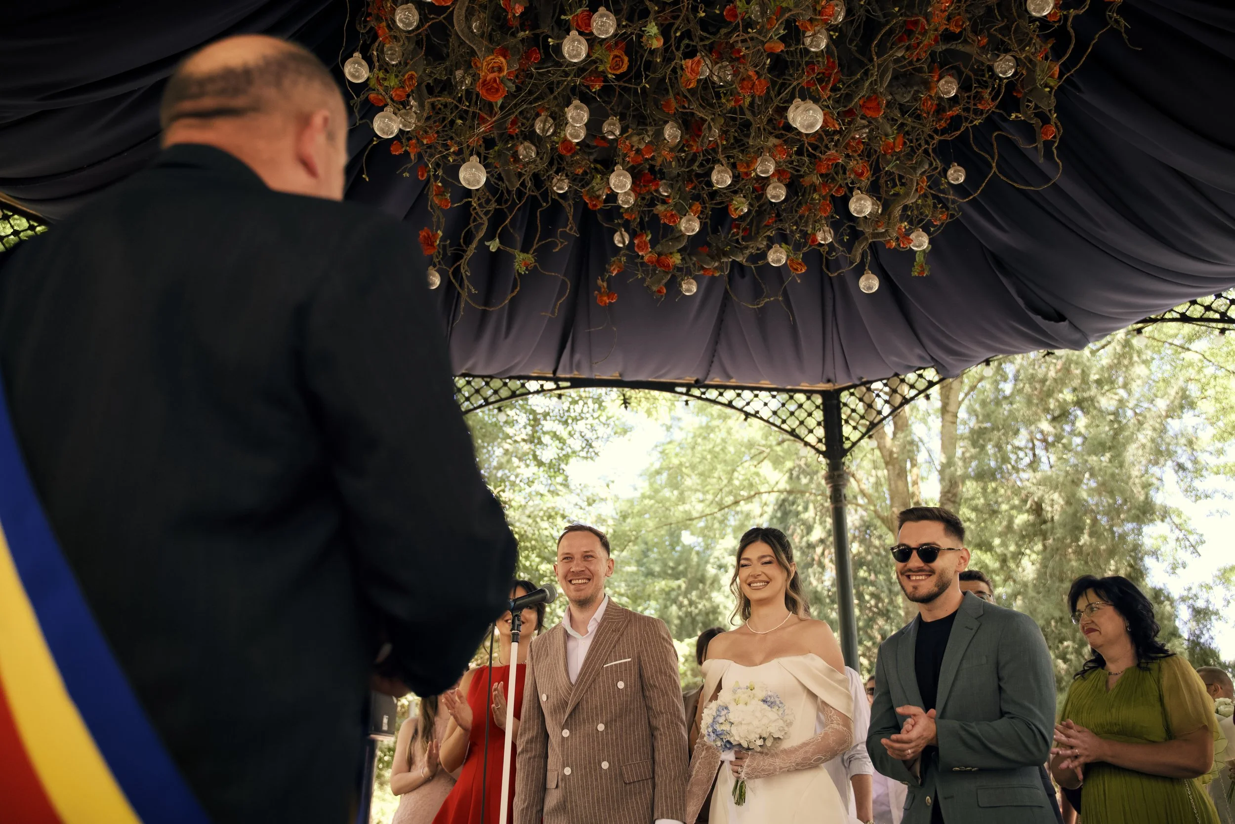 A wedding ceremony outdoors with an officiant in the foreground and groom, bride, and guests smiling under a decorated canopy.