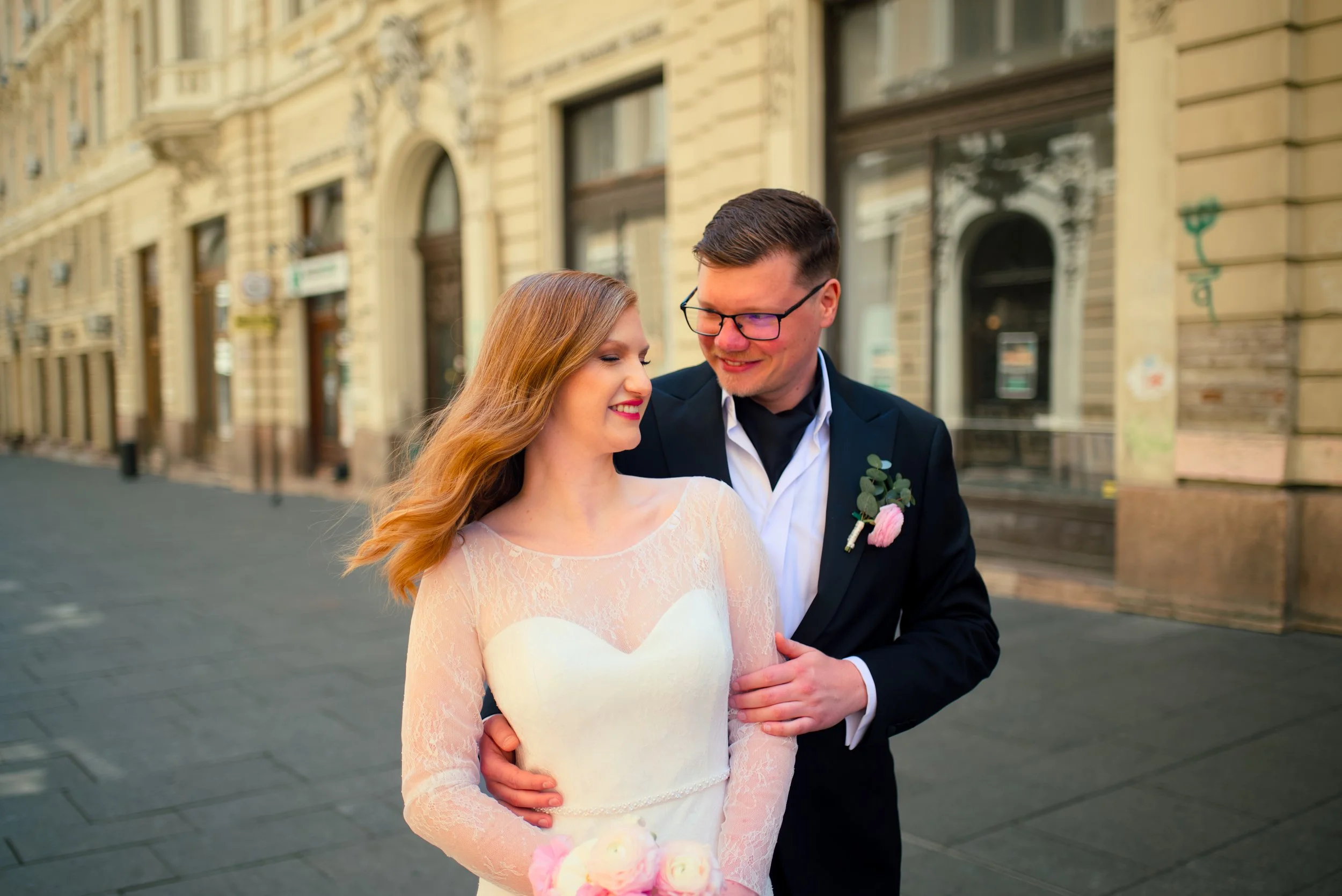 A bride and groom smiling and gazing at each other on a city street, with historic buildings in the background.