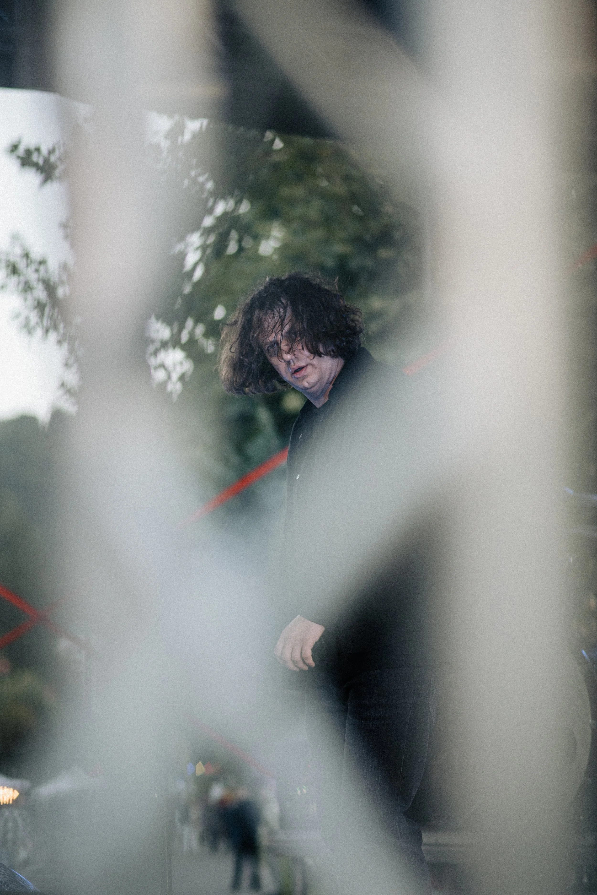 A person with curly hair and glasses wearing a black jacket, captured through an out-of-focus foreground with trees in the background.