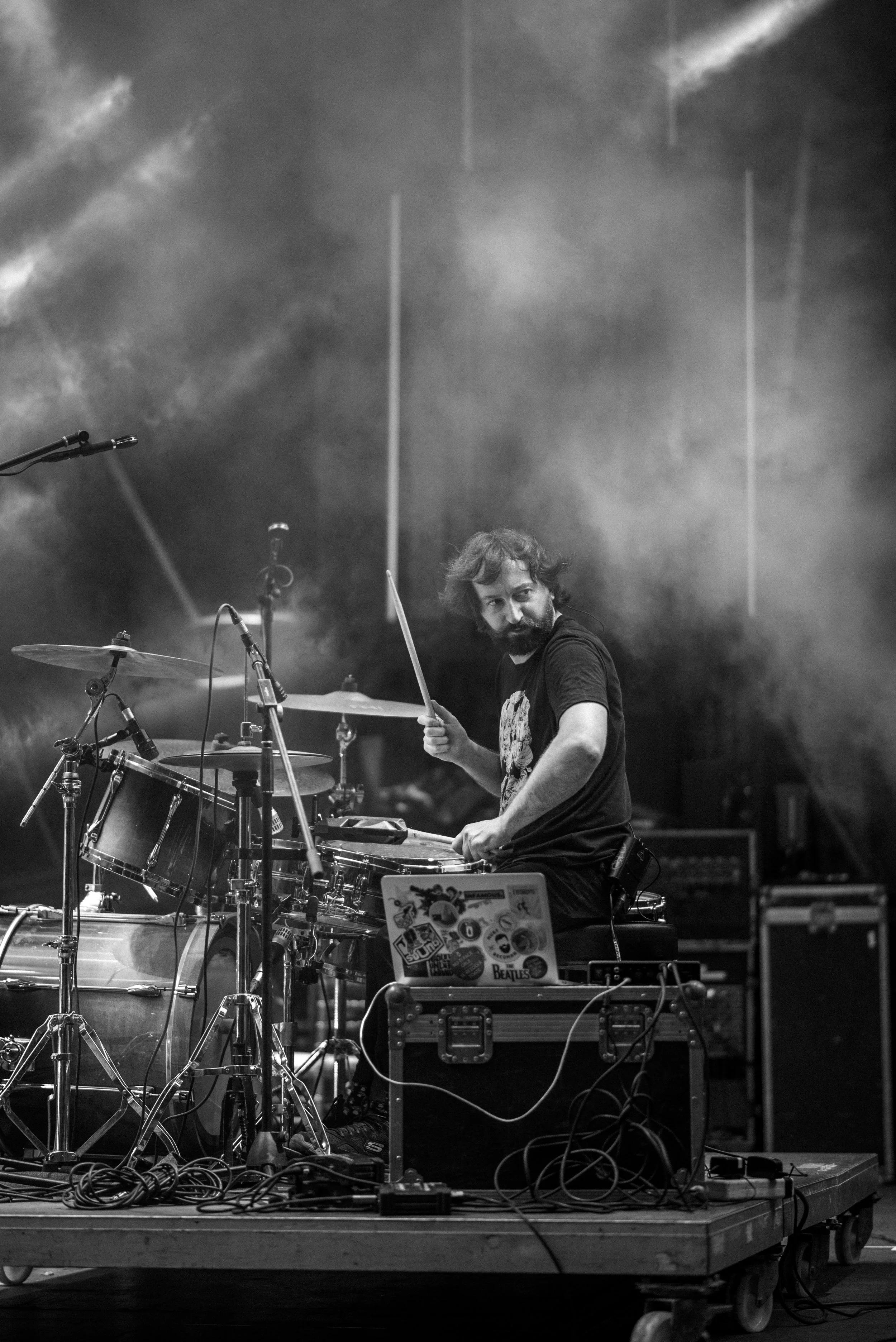 A man with a beard and curly hair playing drums on stage, surrounded by smoke and stage equipment, with a sticker-covered laptop in front of him.
