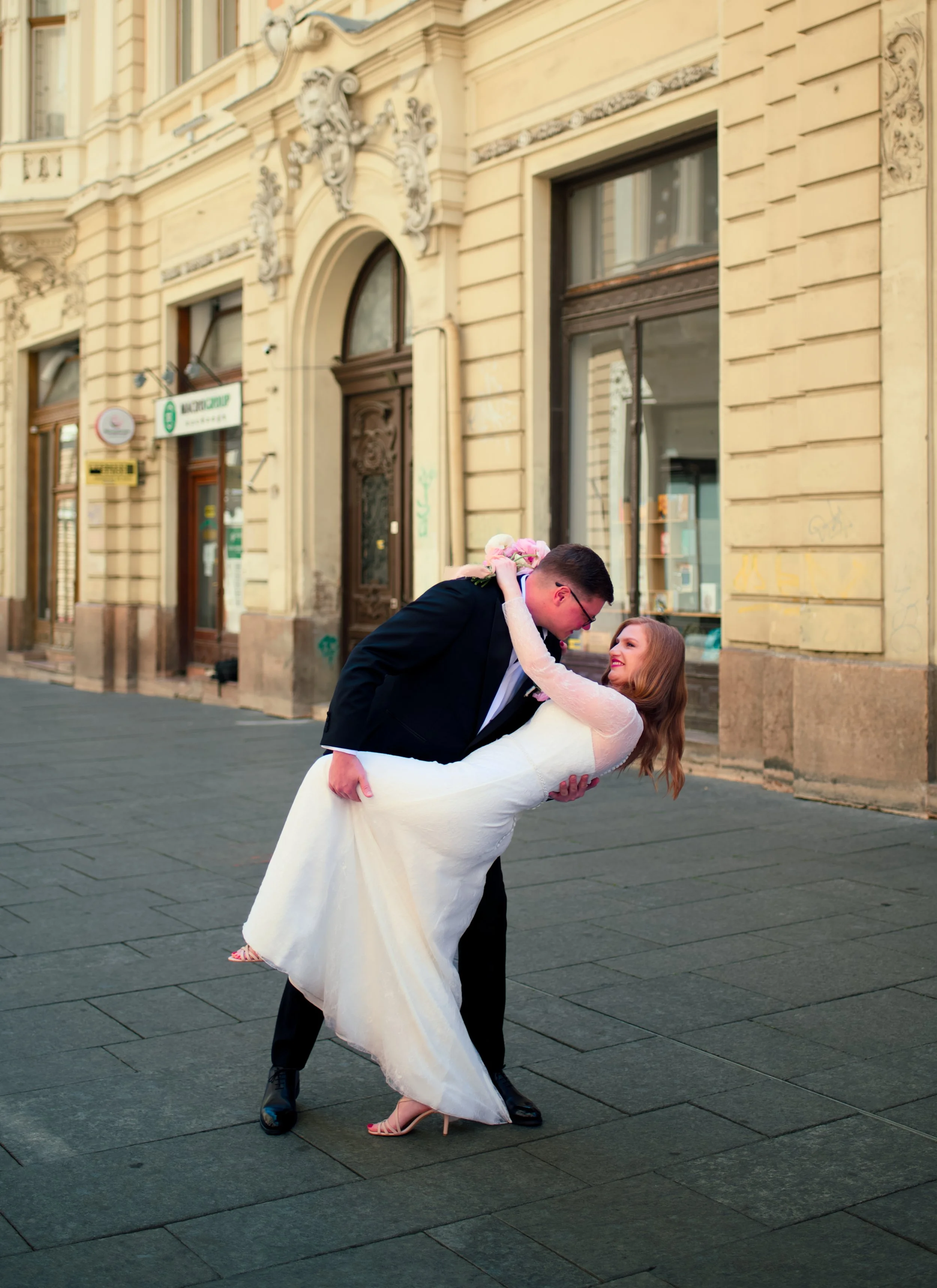 A newlywed couple dancing outside on a city street, with the groom dipping the bride while holding a bouquet, against a backdrop of historic buildings.