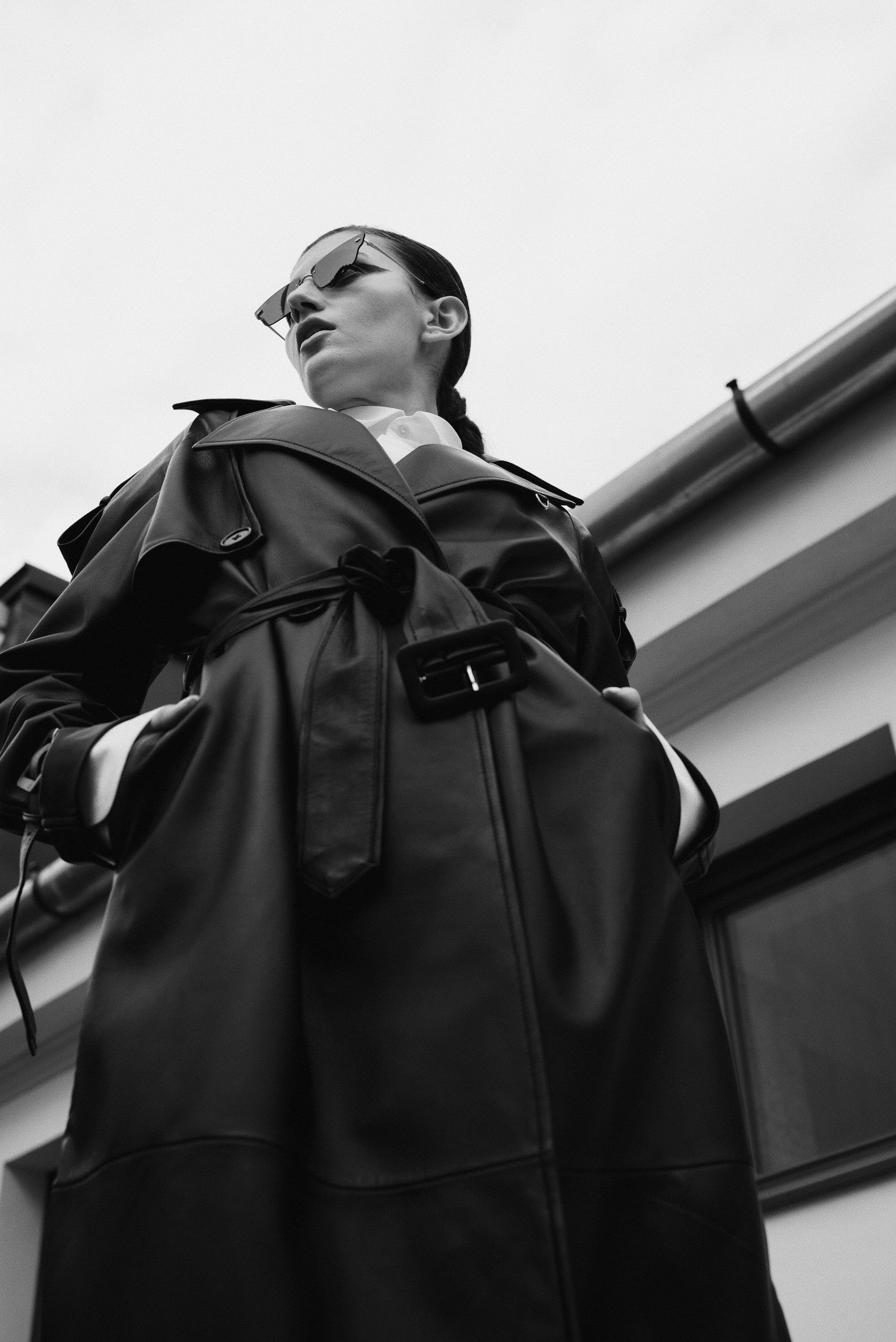 Black and white photo of a woman wearing sunglasses and a trench coat carrying a large bag, looking down from a low angle outdoors.
