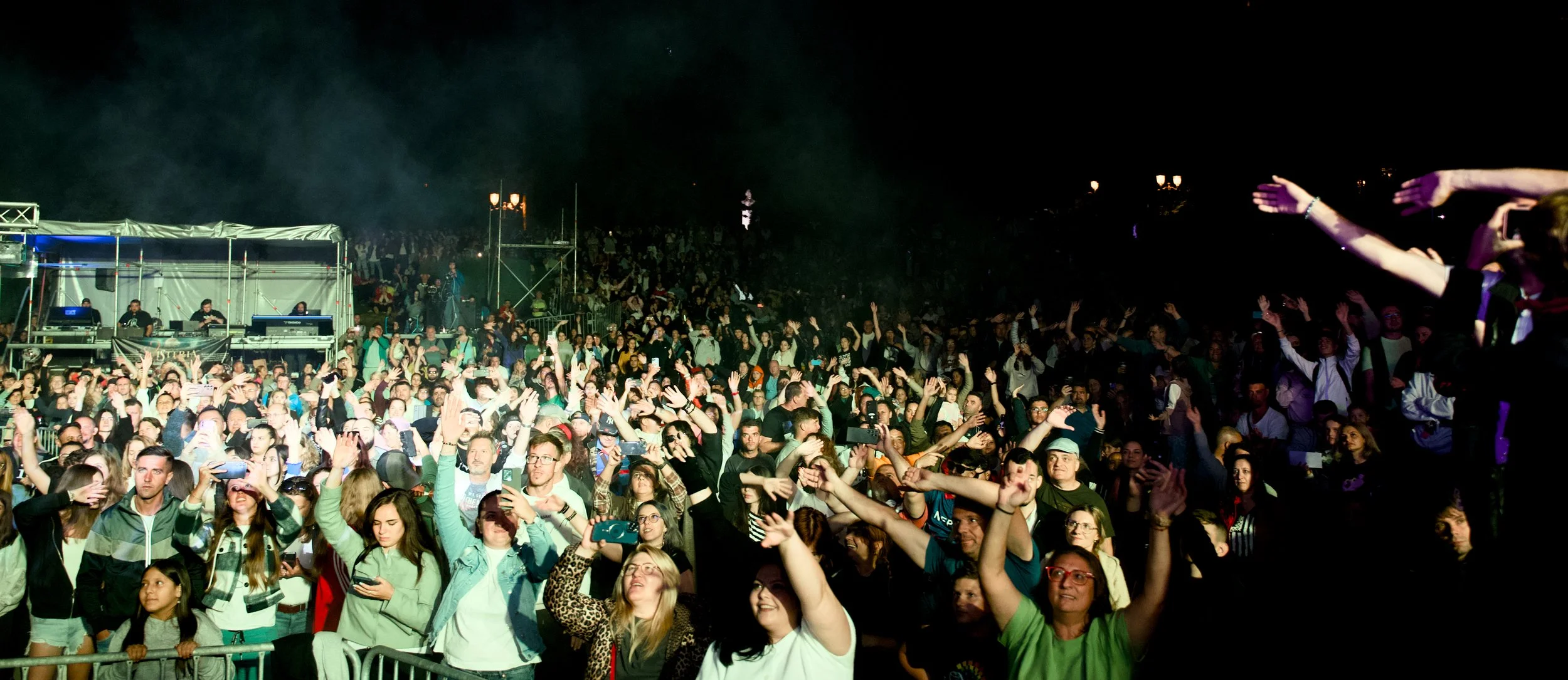 Crowd of people at a night outdoor concert or festival, with some raising their hands and cheering, near a stage with DJ or music equipment.