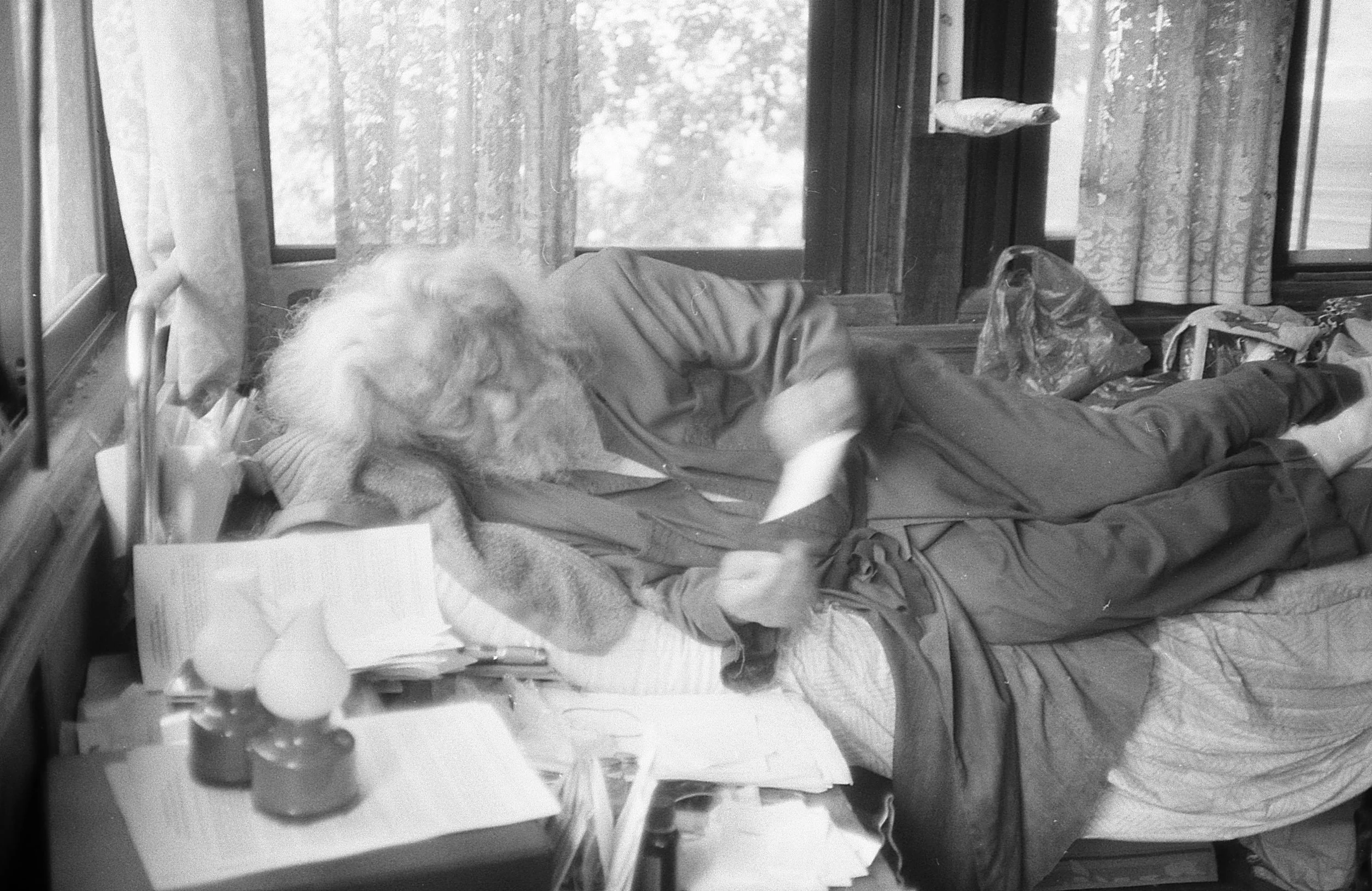 A person with long hair lying on a bed, reaching for items on a nightstand next to a window.