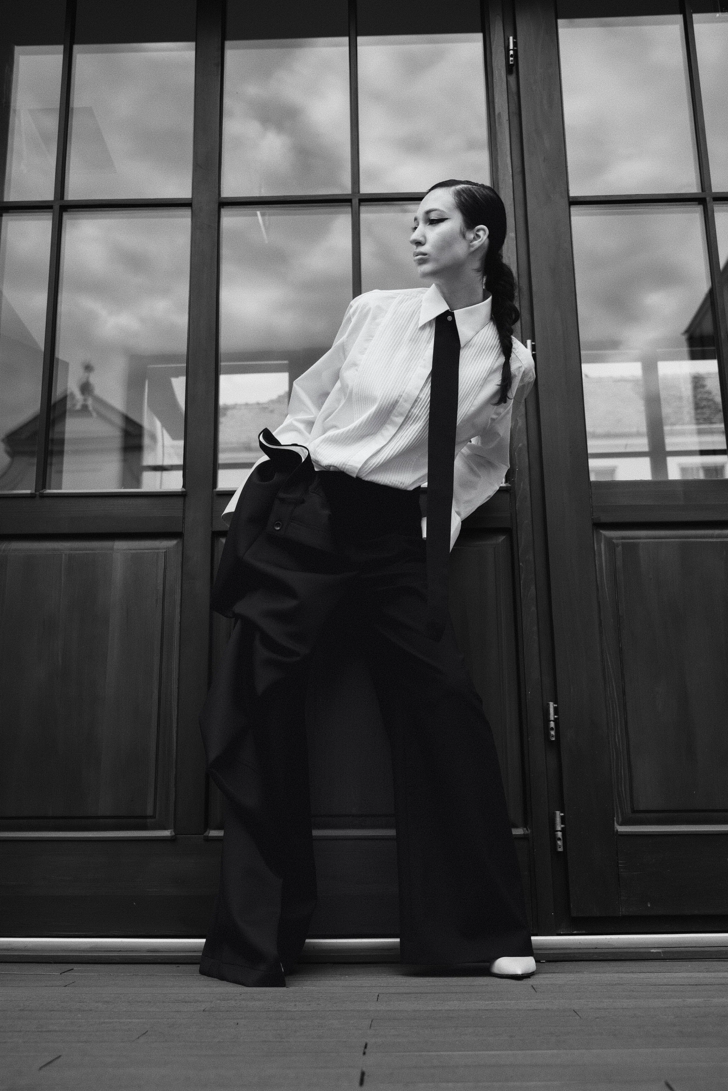 A woman in a white shirt and black wide-leg pants leaning against a wooden and glass door, looking away with a serious expression, in black and white.