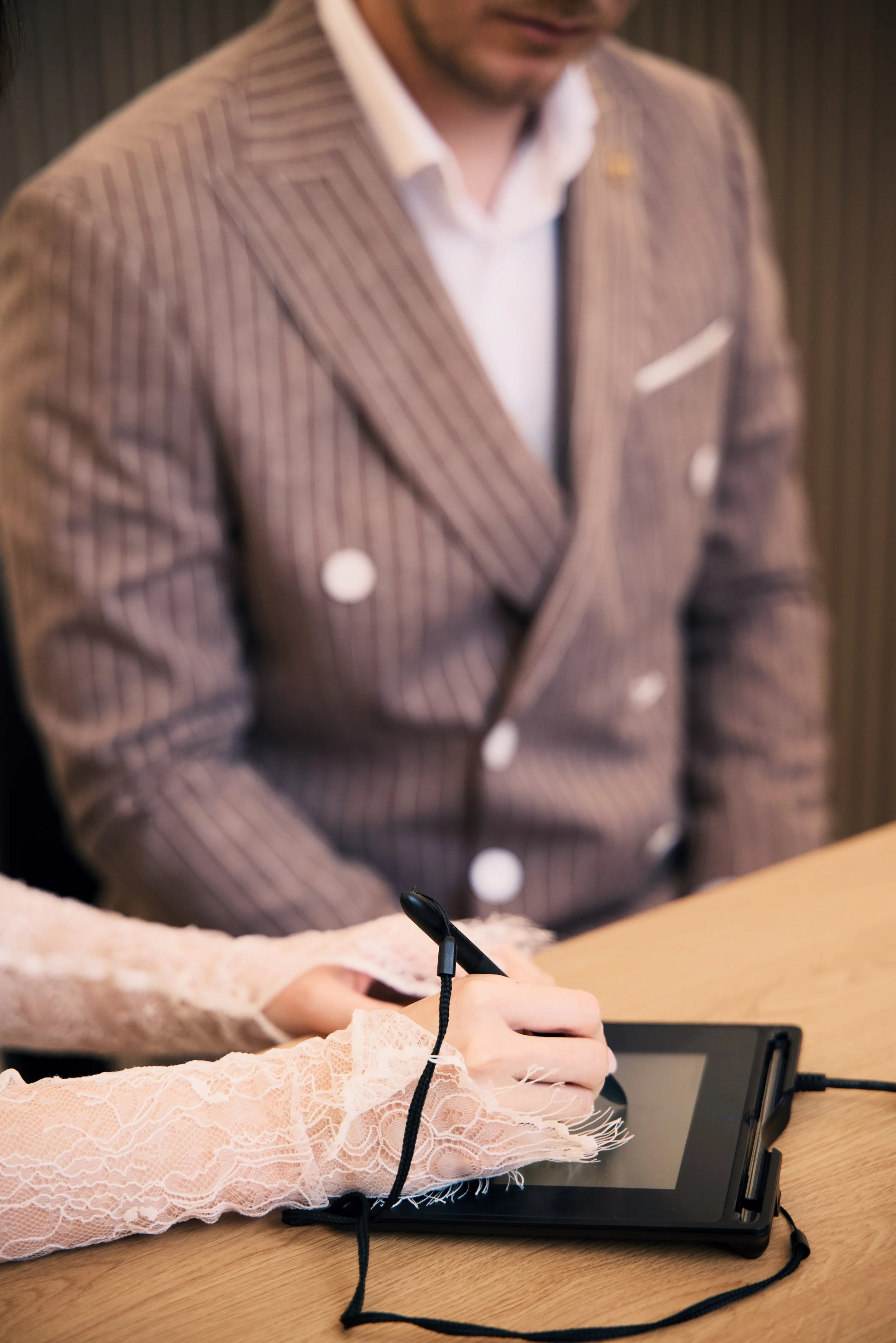 Person in a beige lace sleeve using a black drawing tablet with a stylus on a wooden table, man in a striped suit in the background.