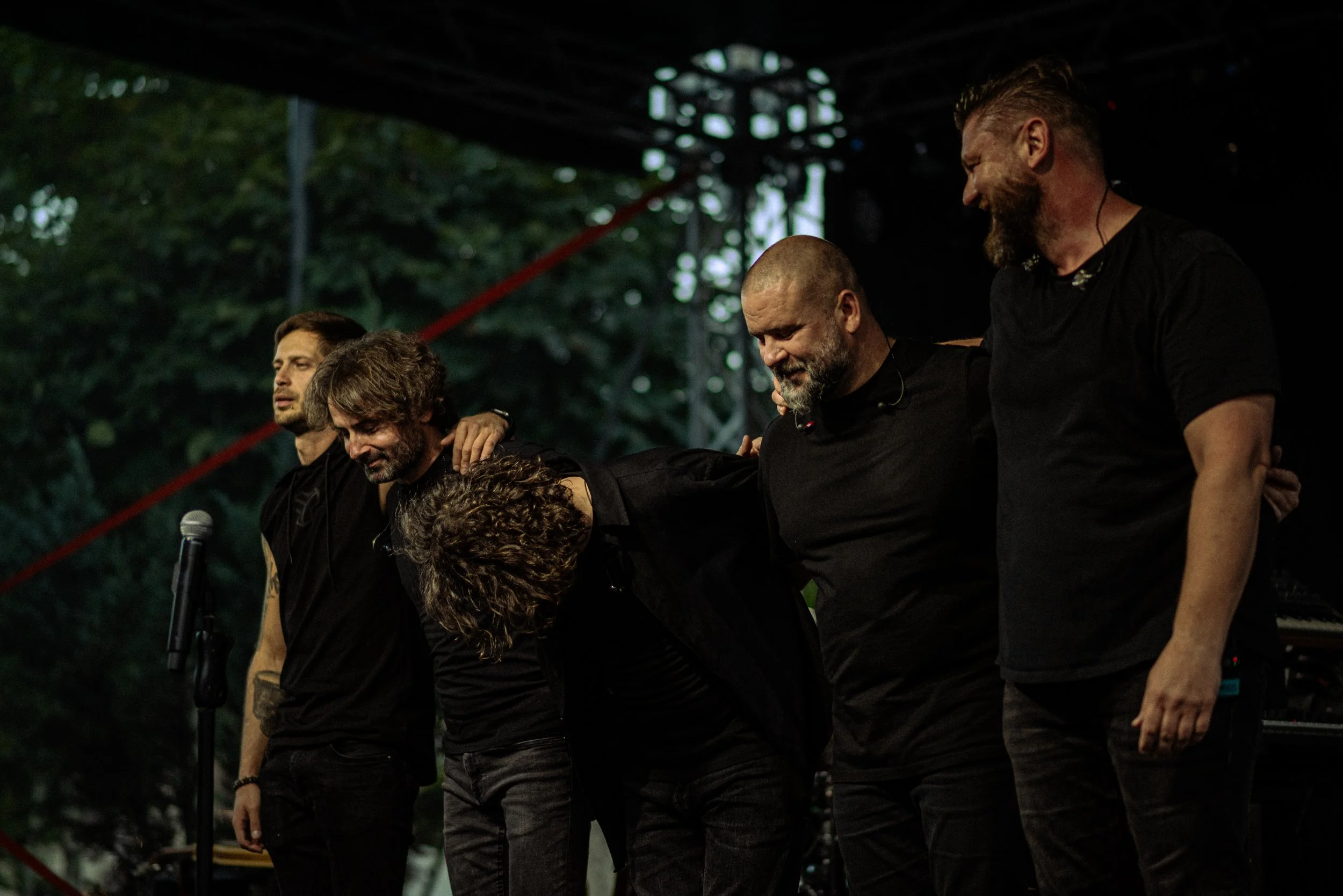 Five men in black shirts standing side by side on a stage, with their arms around each other's shoulders, bowing and smiling.