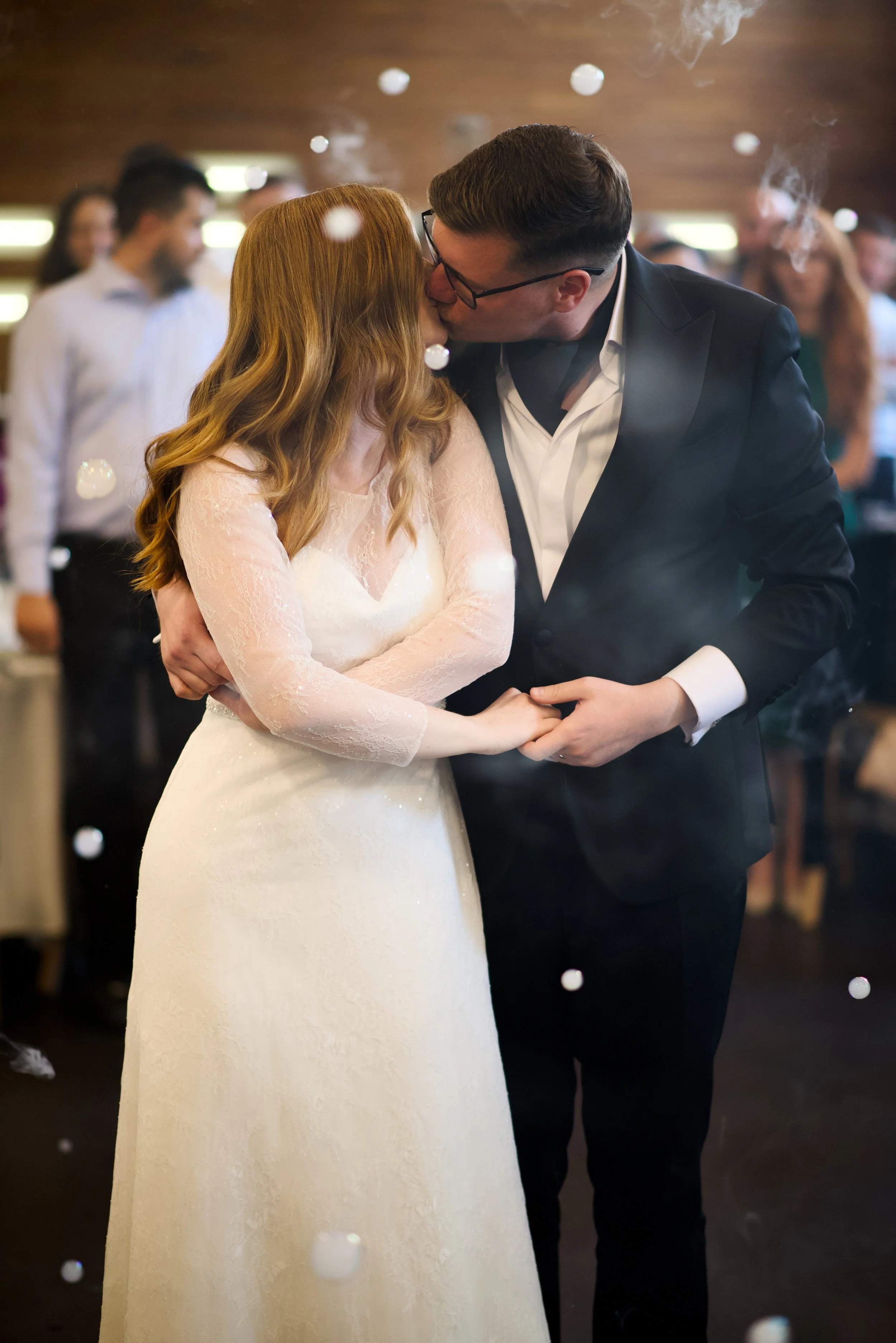 A bride and groom kiss during their wedding reception, holding hands and embracing, with guests in the background.
