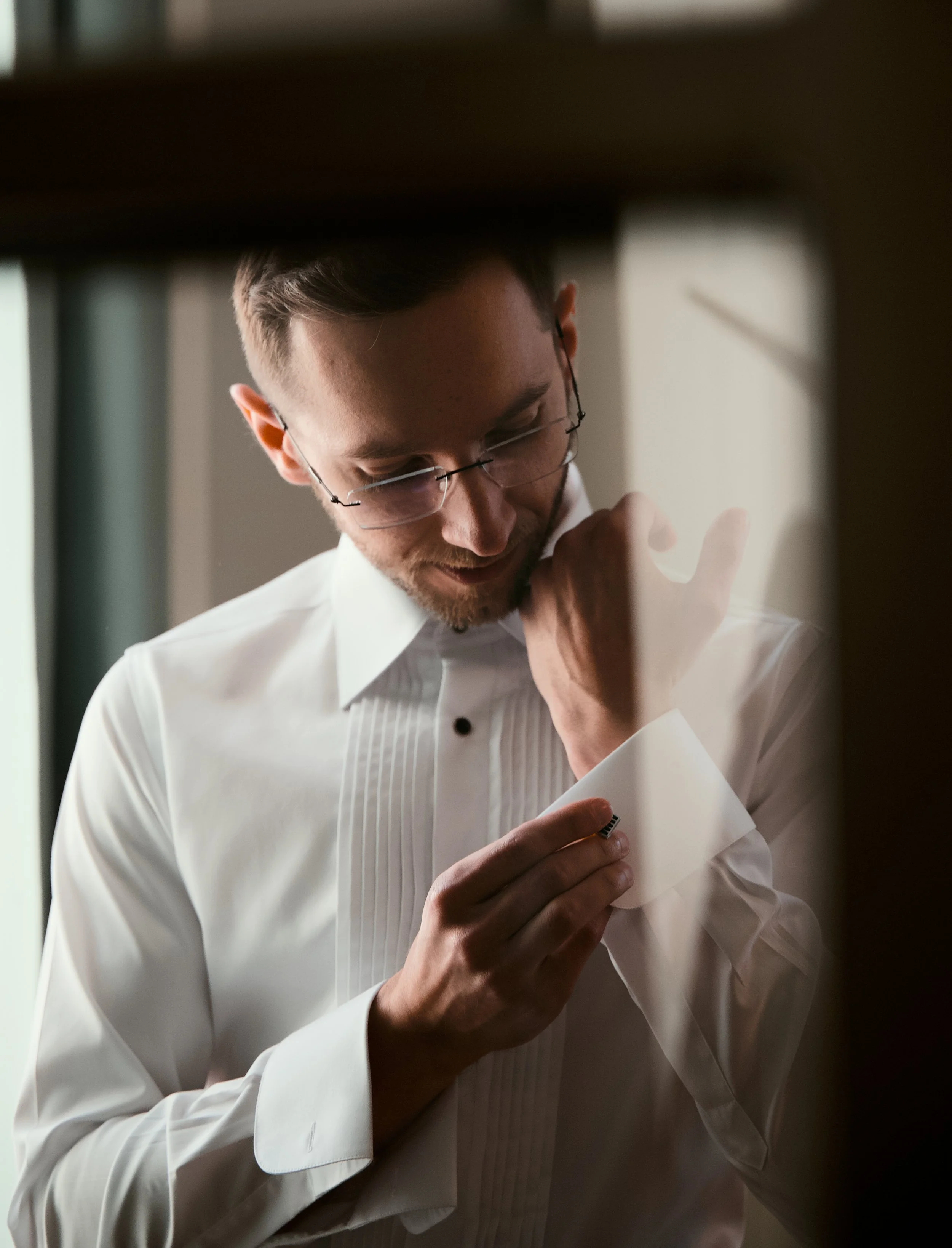Man in white shirt with black buttons and glasses looking down and holding a cufflink or jewelry piece near his wrist.