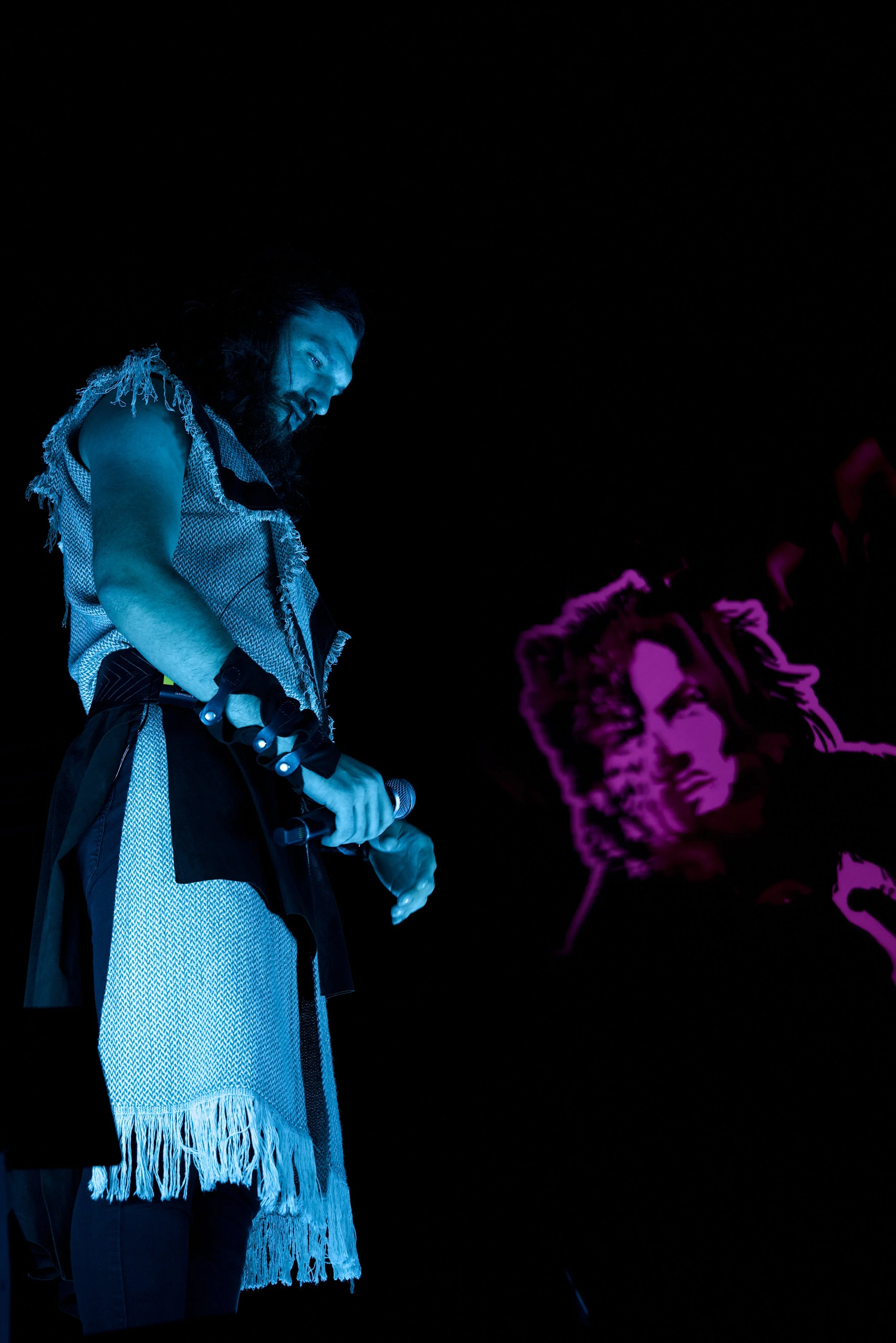 Performer with long dark hair and a beard, wearing a sleeveless, fringed outfit, holding a microphone on stage with a purple-lit portrait of a woman projected in the background.