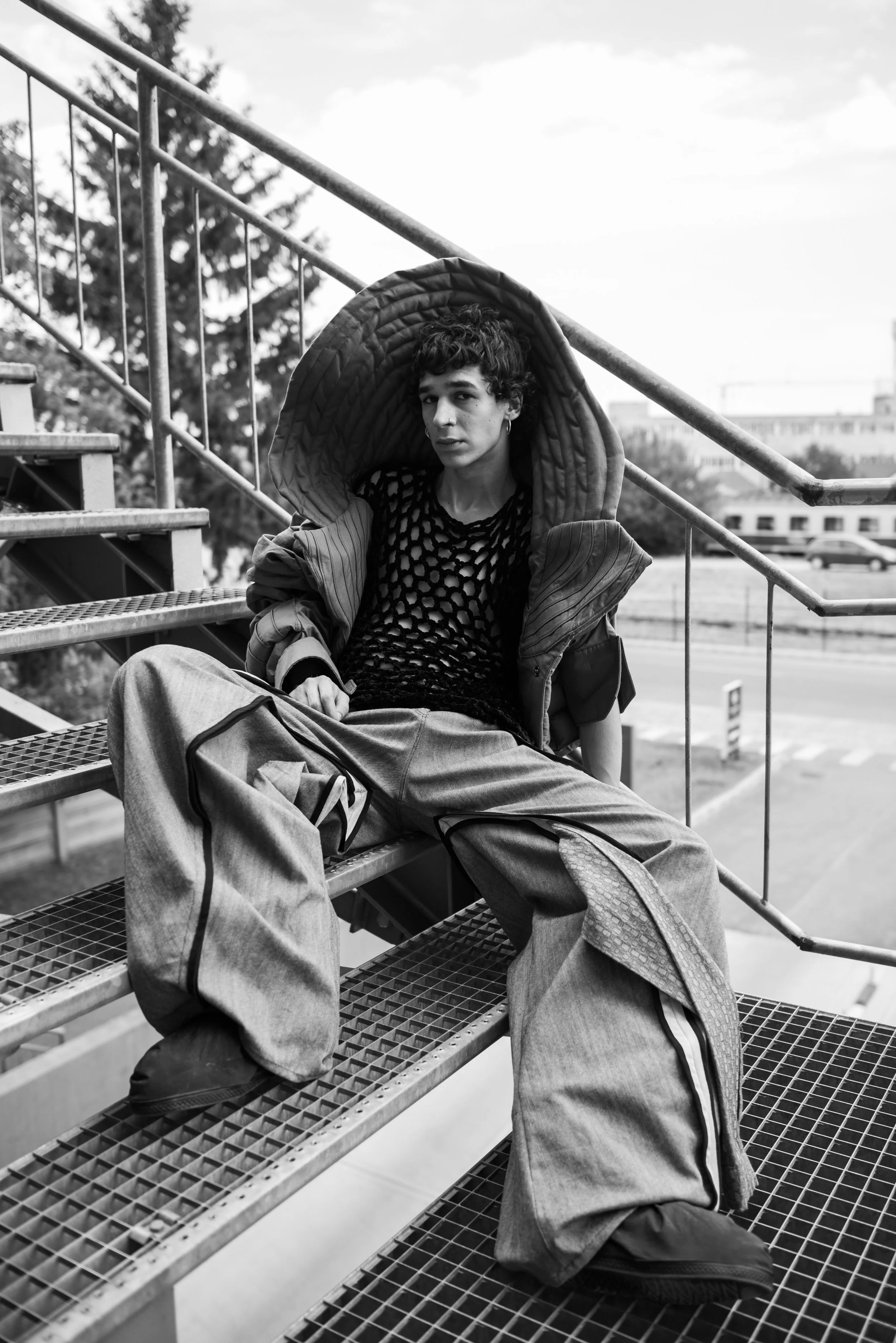 A young man with curly hair sitting on metal outdoor stairs, wearing baggy pants, a black mesh shirt, and a large jacket with a hood. The photo is in black and white, and he appears to be looking at the camera with a serious expression.