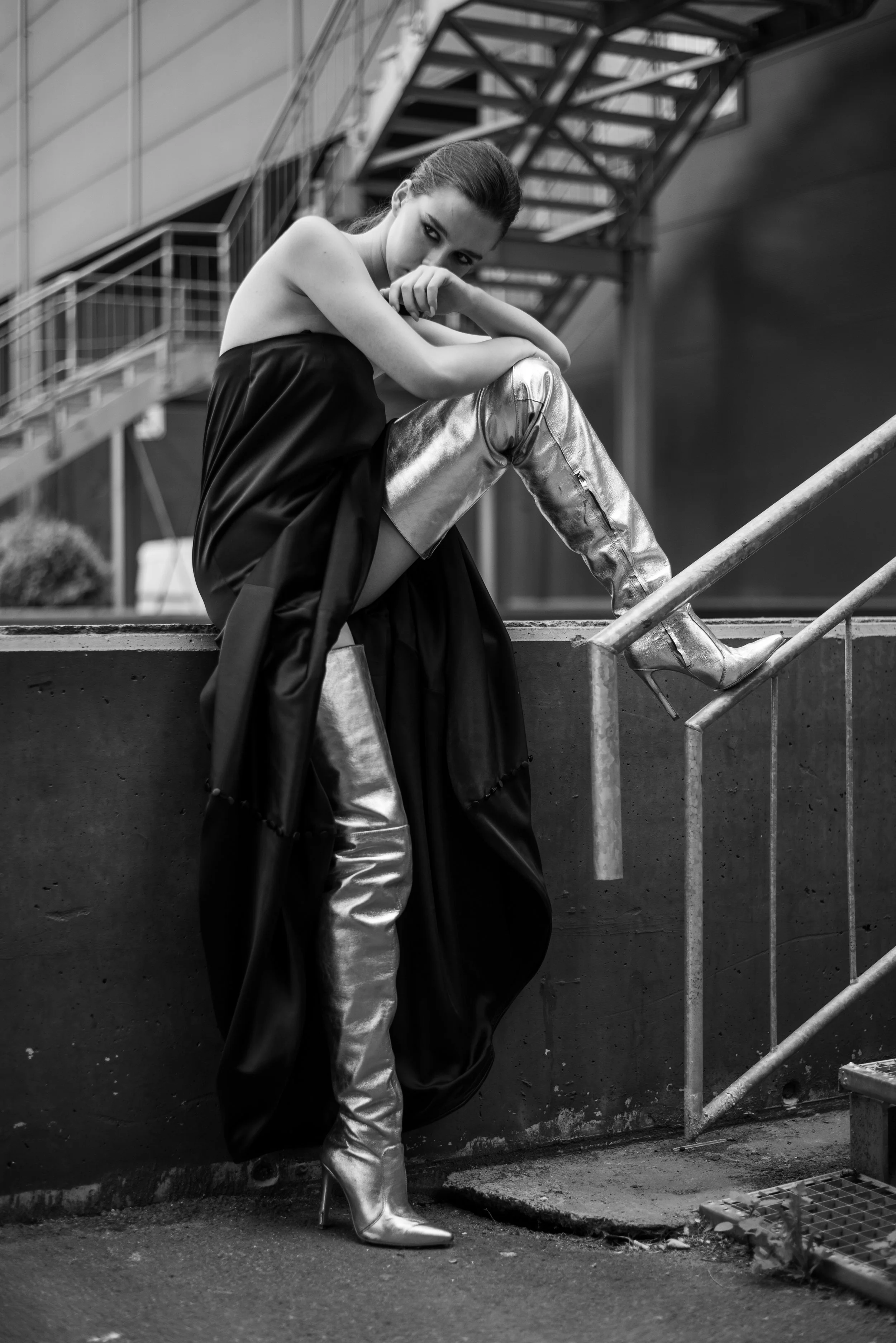 A black and white photo of a woman sitting on a ledge, wearing shiny, high-heeled knee-high boots, metallic pants, and a flowing dark dress, with a modern industrial background.