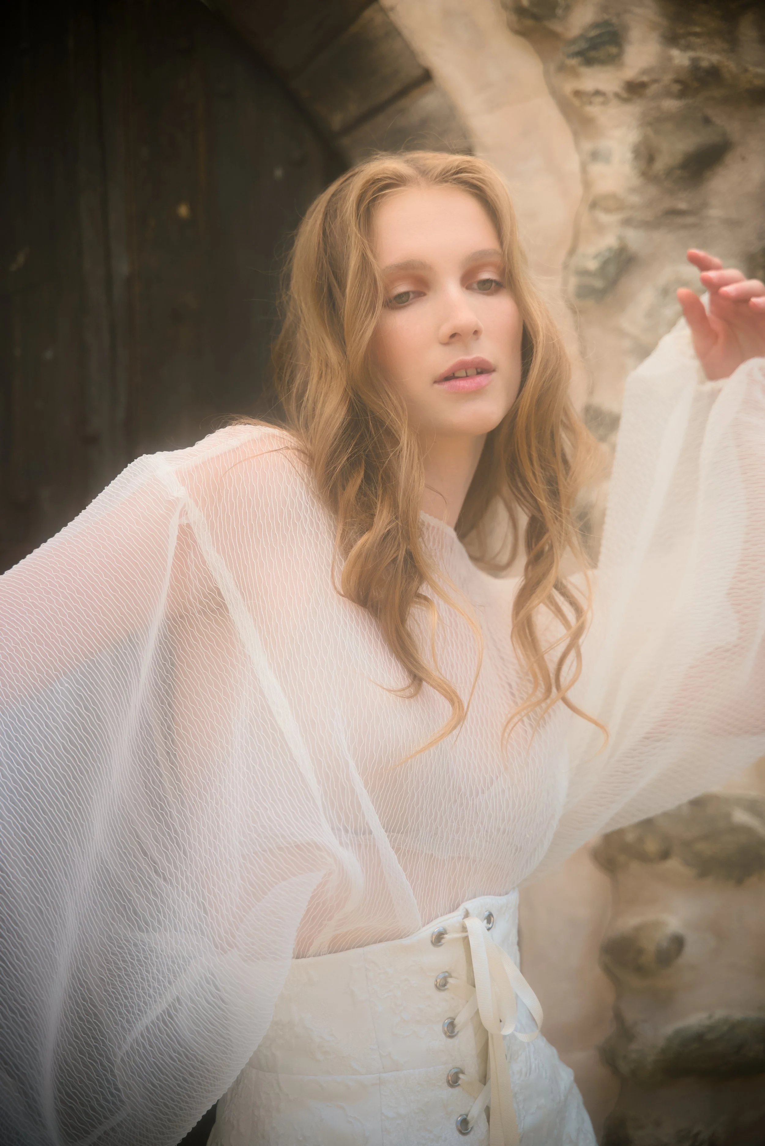 A woman with wavy light brown hair wearing a sheer white top and white high-waisted shorts standing against a stone and wooden background.