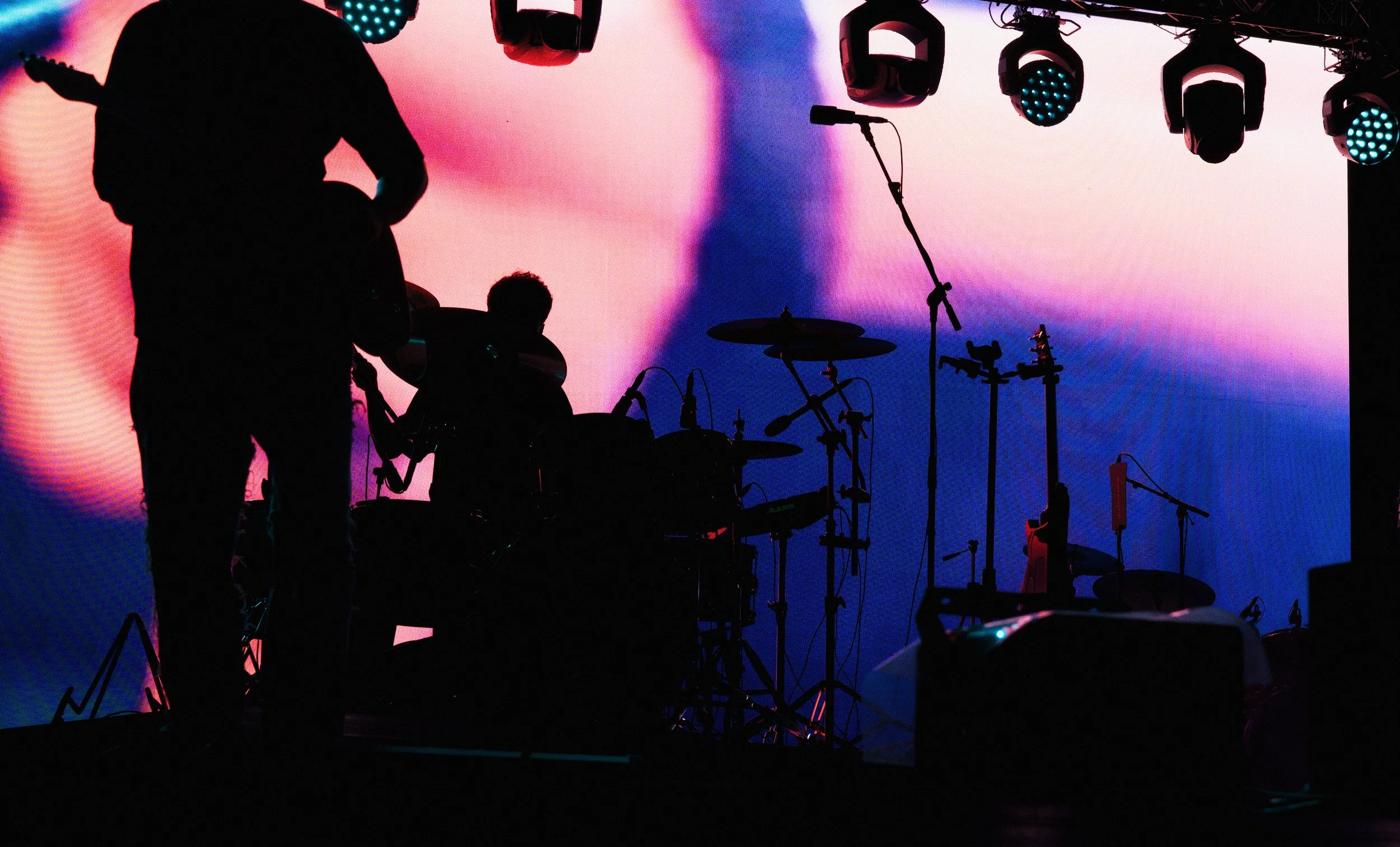 Silhouettes of musicians with drums and microphones on stage, illuminated by colorful stage lights and a vibrant background screen.