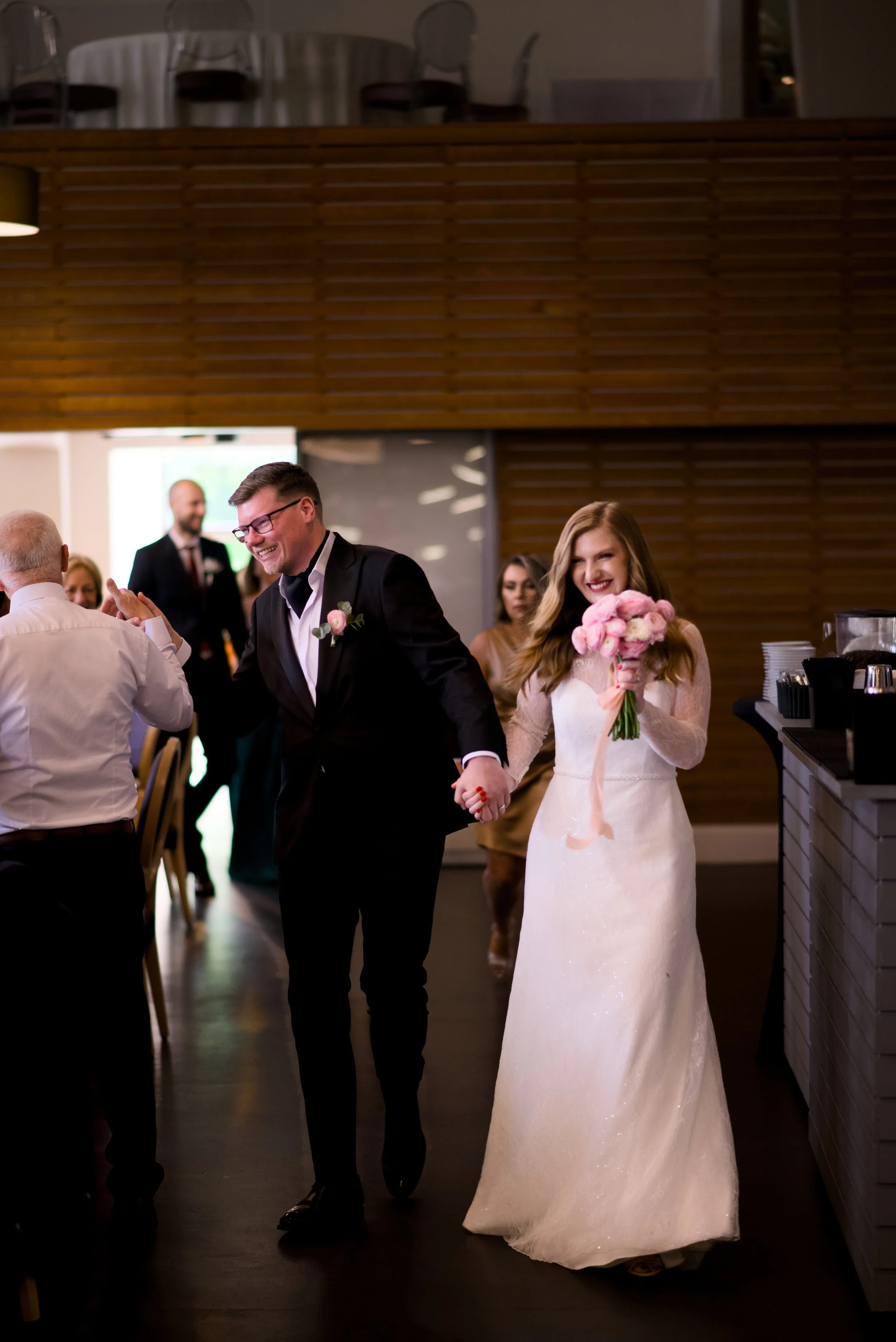 A newlywed couple holding hands and smiling at each other at a wedding reception. The bride is holding a bouquet of pink flowers, and the groom is dressed in a black tuxedo with a pink boutonniere. Guests are visible in the background.