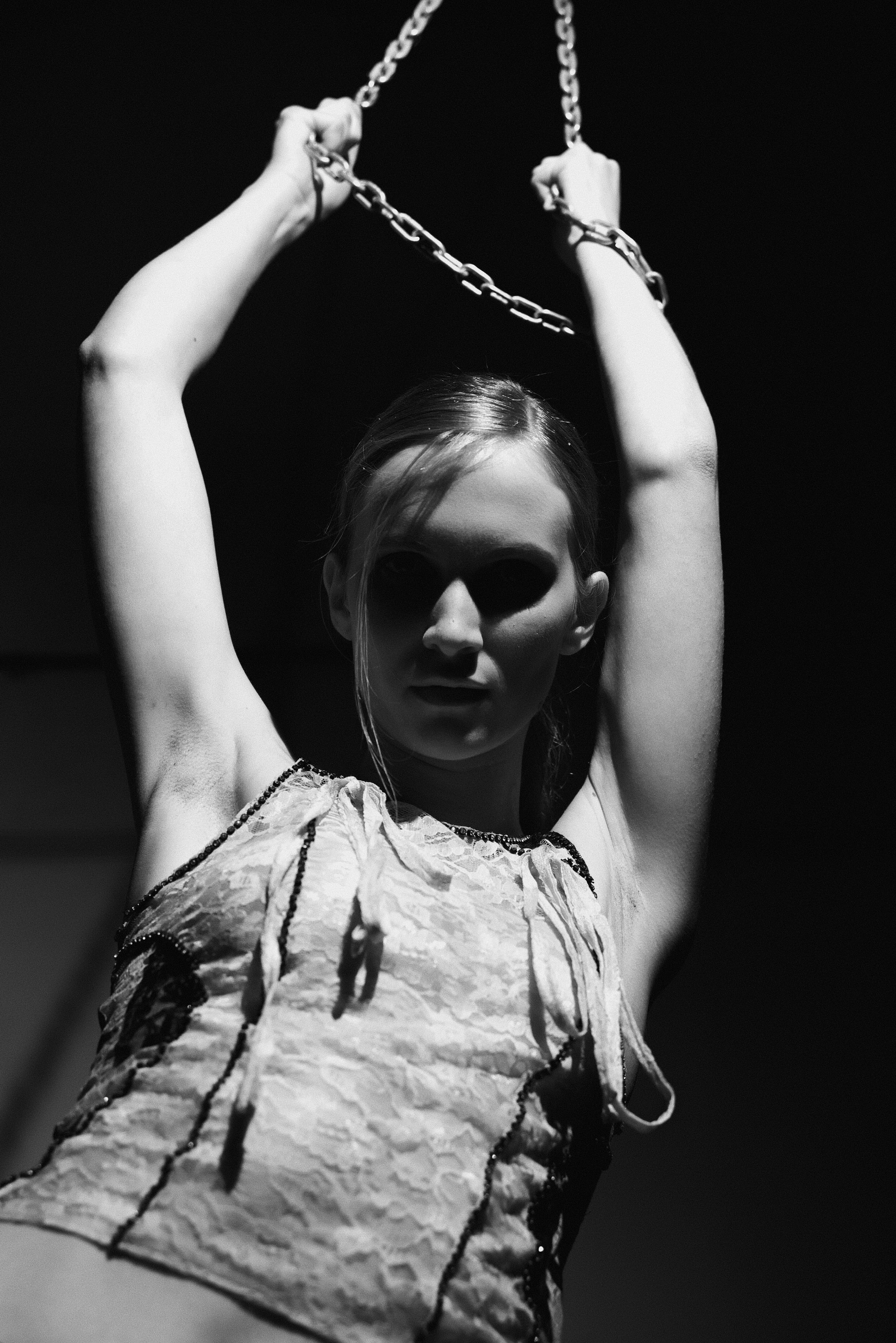 A woman with arms raised holding a chained object above her head, with dramatic lighting casting shadows on her face, in a black and white photograph.