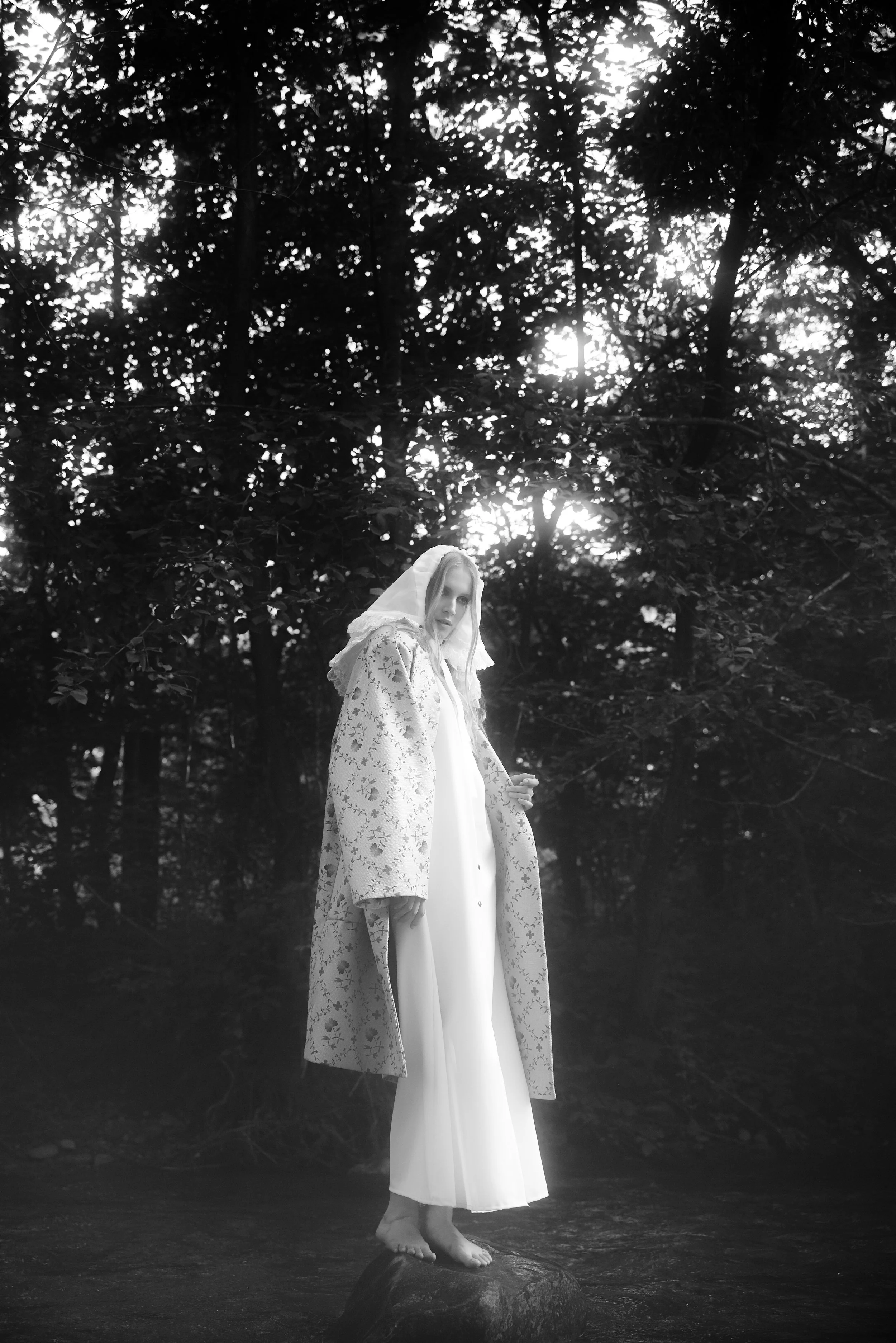 A woman standing barefoot on a rock in a forest with sunlight filtering through the trees, wearing a white dress and a patterned coat with a hood.