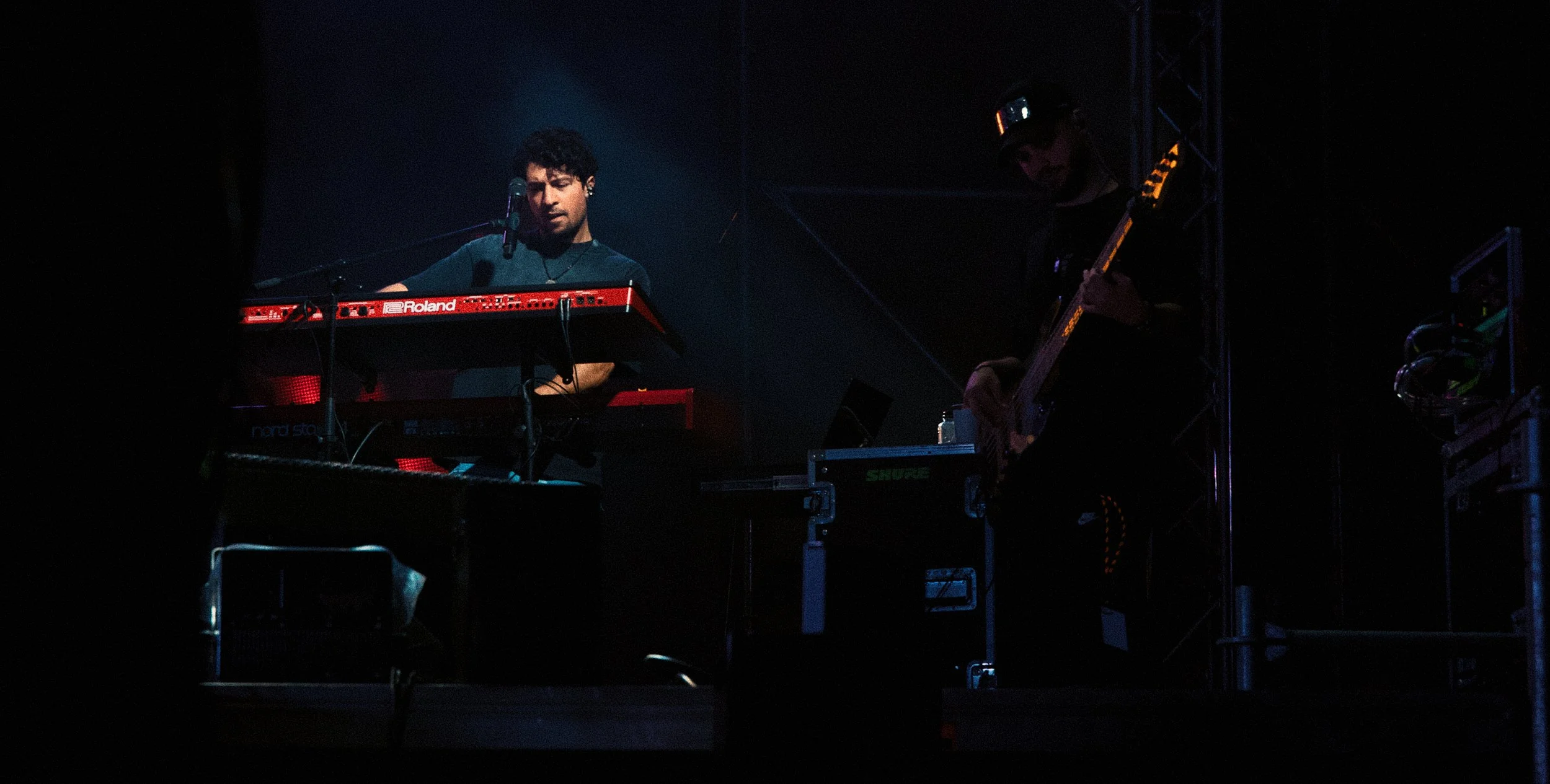 Two musicians performing on stage: one playing keyboards and the other playing guitar, in a dark setting with stage equipment visible.
