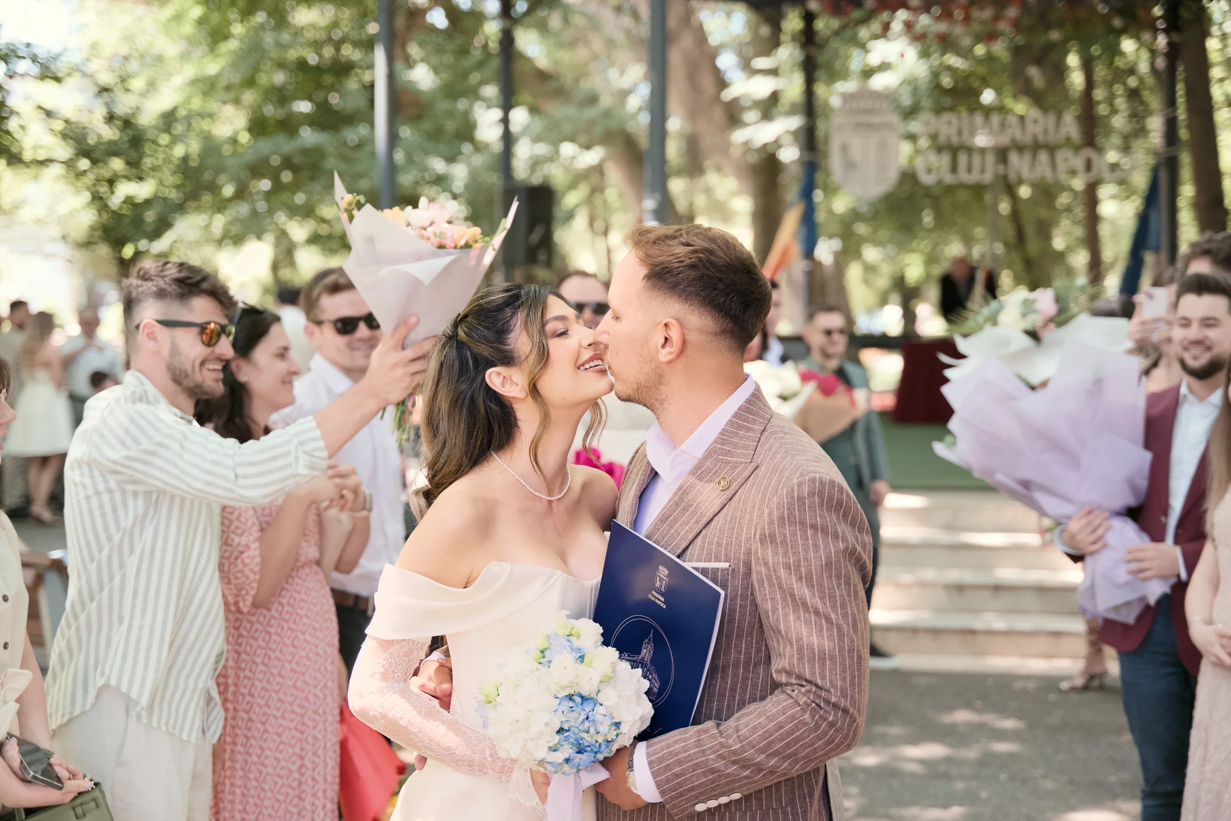 A newlywed couple sharing a kiss at their outdoor wedding, surrounded by friends holding bouquets of flowers, in a park setting with trees and a sign in the background.