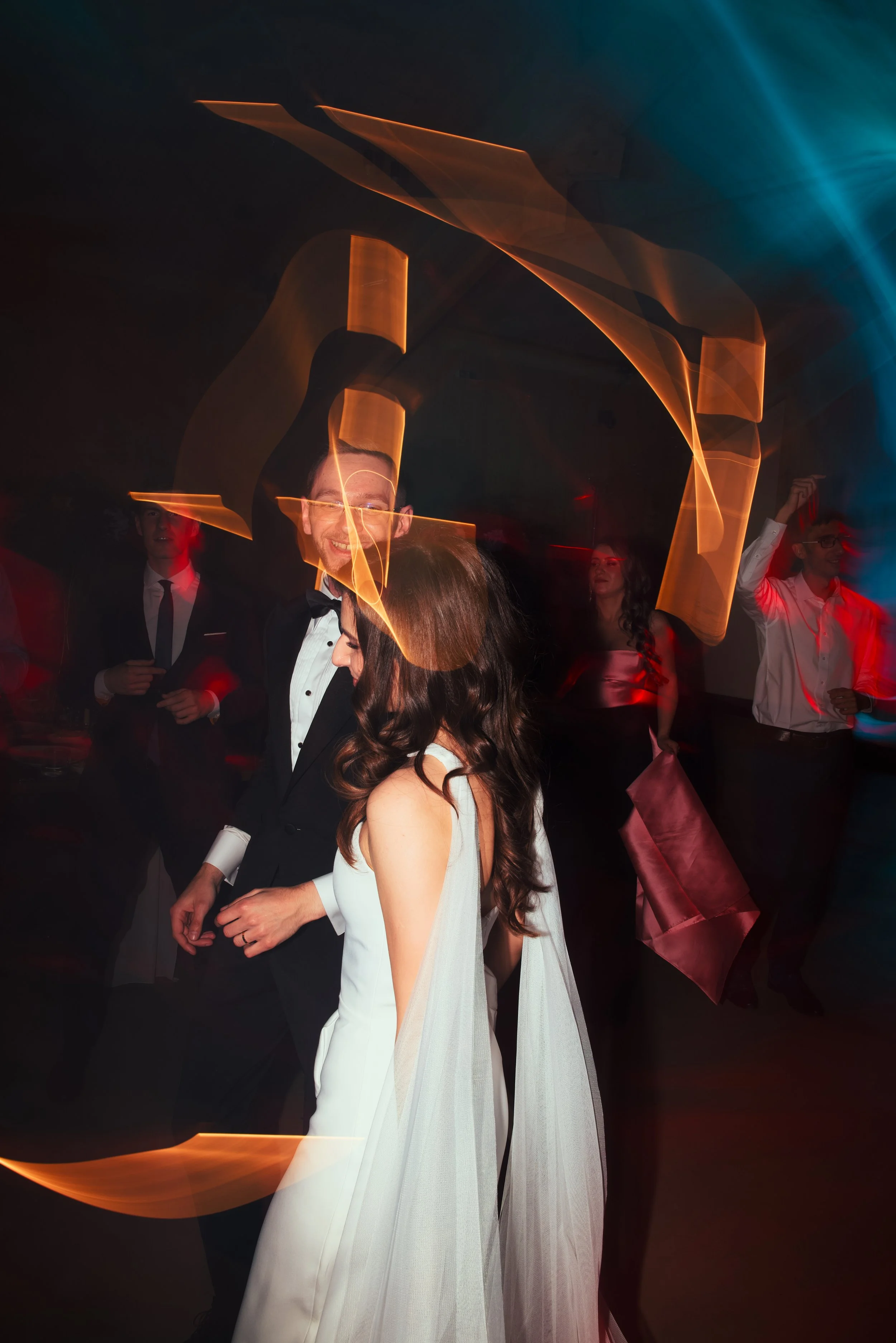 A bride and groom dancing at their wedding reception with colorful light trails and other guests dancing in the background.