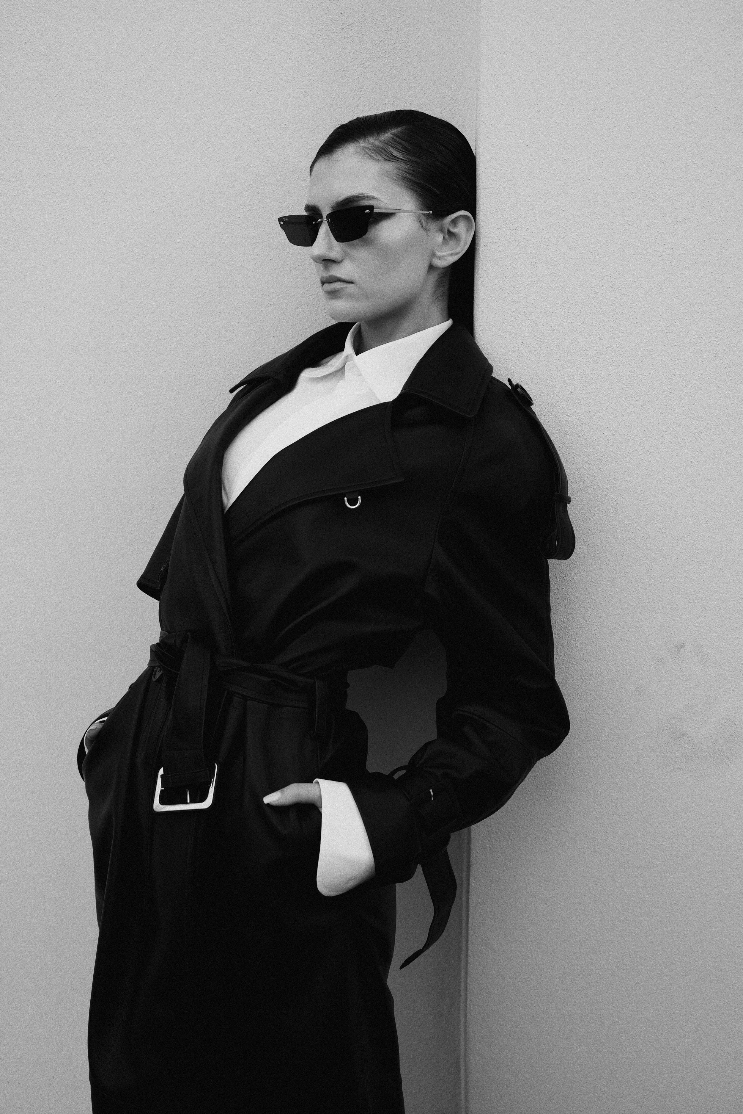 A woman wearing sunglasses, a white shirt, and a black leather jacket, leaning against a wall with her hands in her pockets.