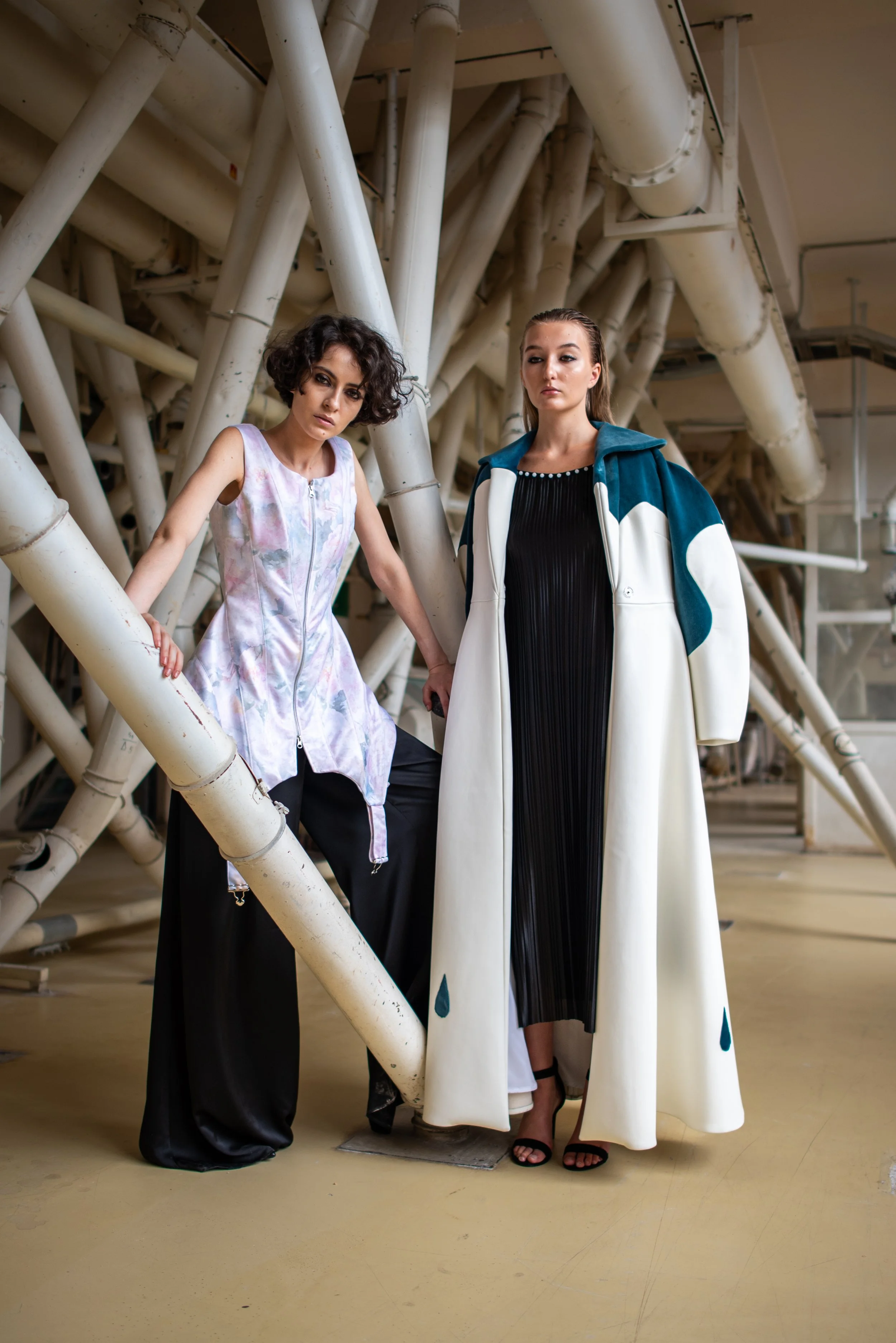 Two women in fashionable clothing standing among industrial pipes.