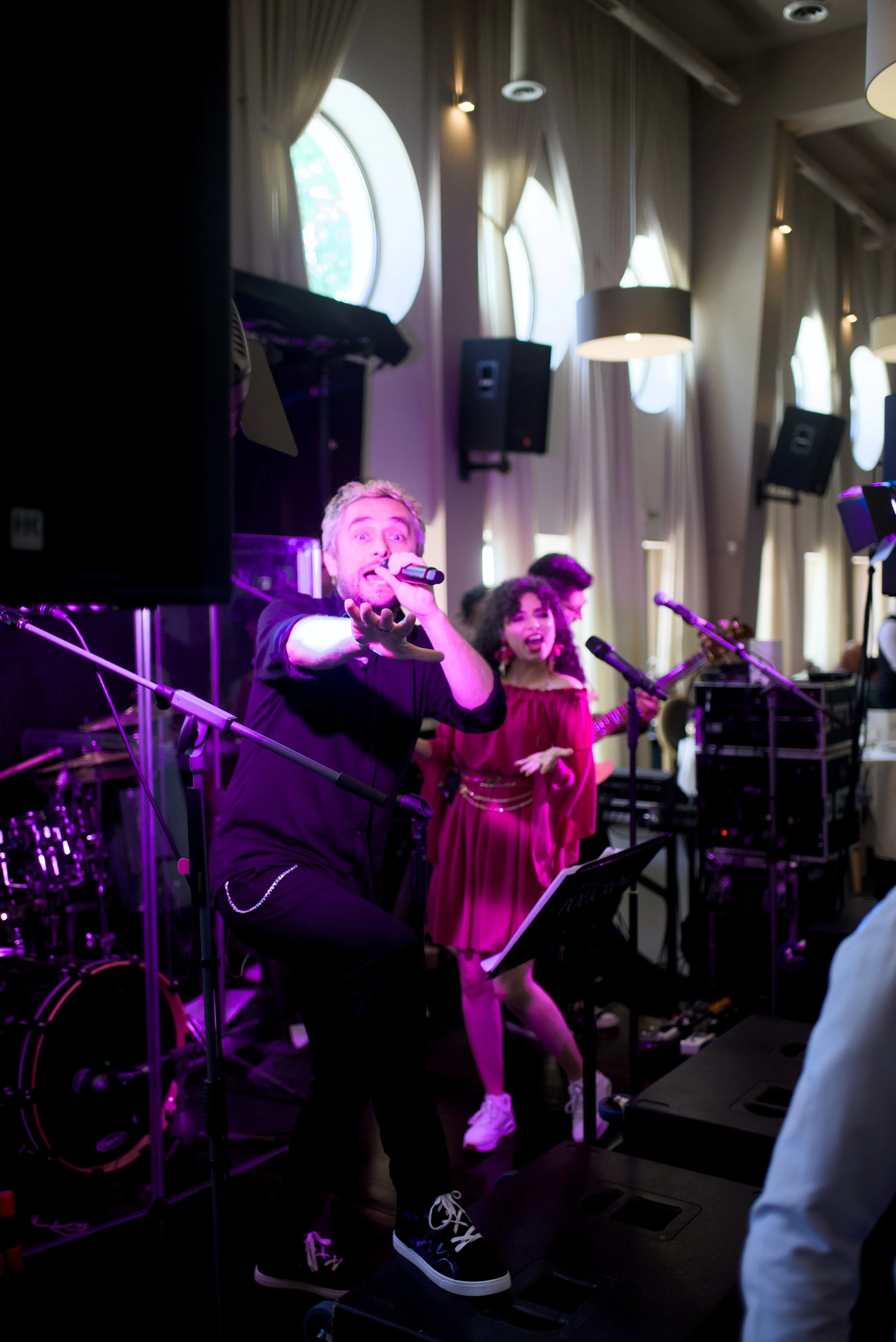 Musicians performing with microphones, a drummer in the background, in a room with large windows and curtains, under purple stage lighting.