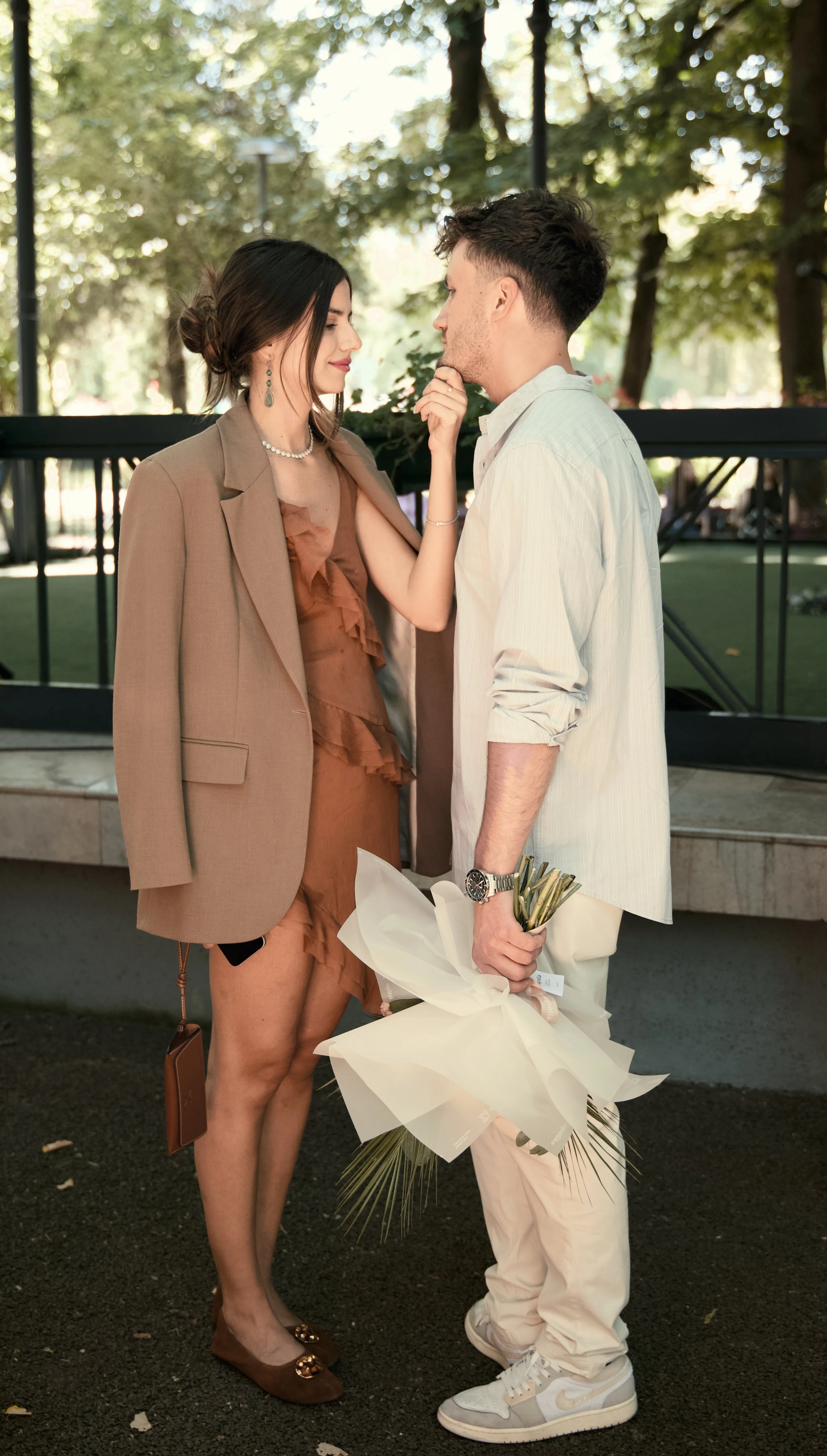 A man and woman stand close in a park, gazing at each other with the woman’s hand touching the man’s chin. The man holds a bouquet of flowers wrapped in white paper. The woman wears a brown dress and a beige blazer, while the man wears a white shirt