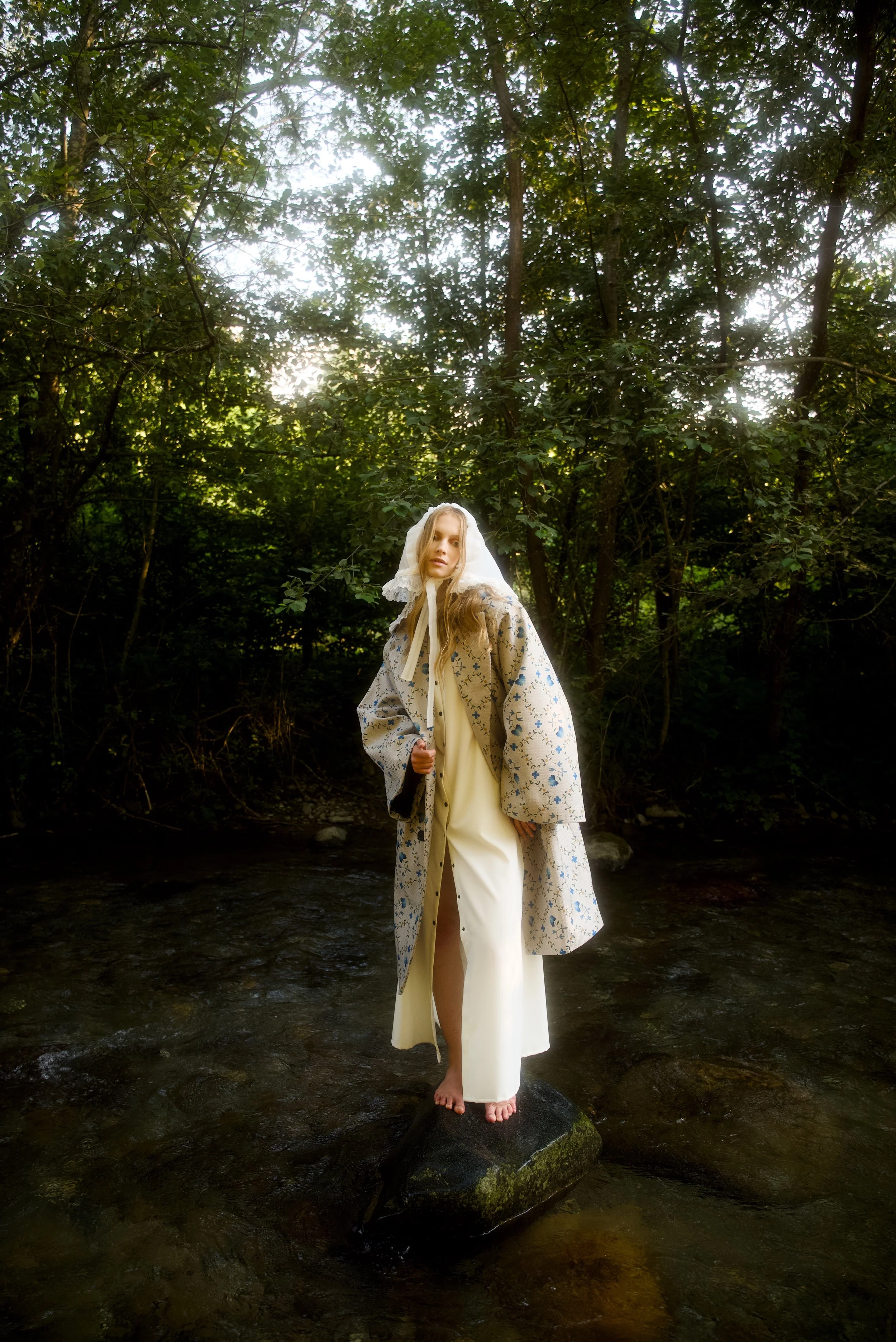 A woman with long blonde hair, wearing vintage-style clothing and a lace head covering, standing barefoot on a rock in a shallow creek surrounded by dense green trees.