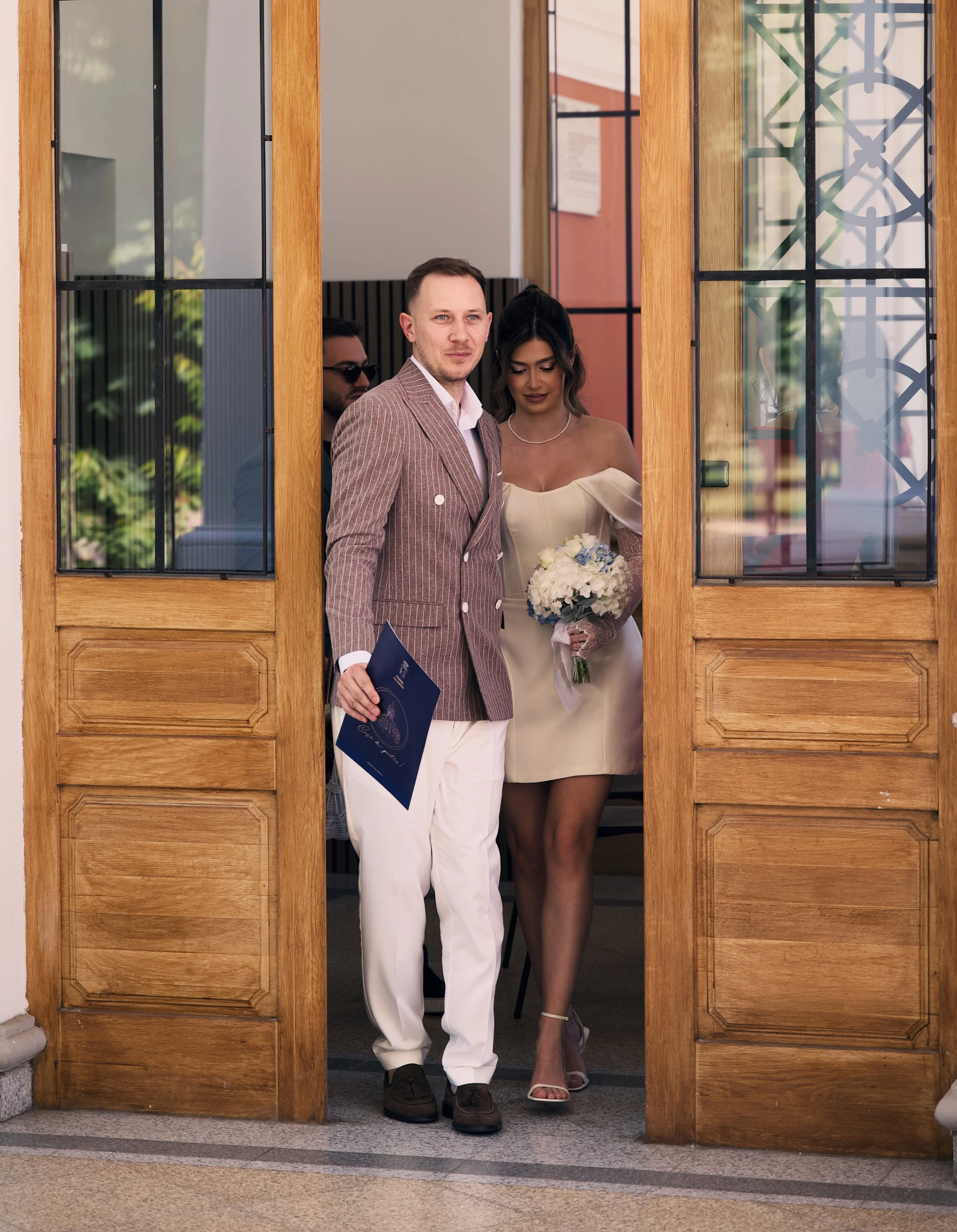 A couple walking out of a building, dressed in formal attire, with the woman holding a bouquet of white flowers and the man holding a folder.