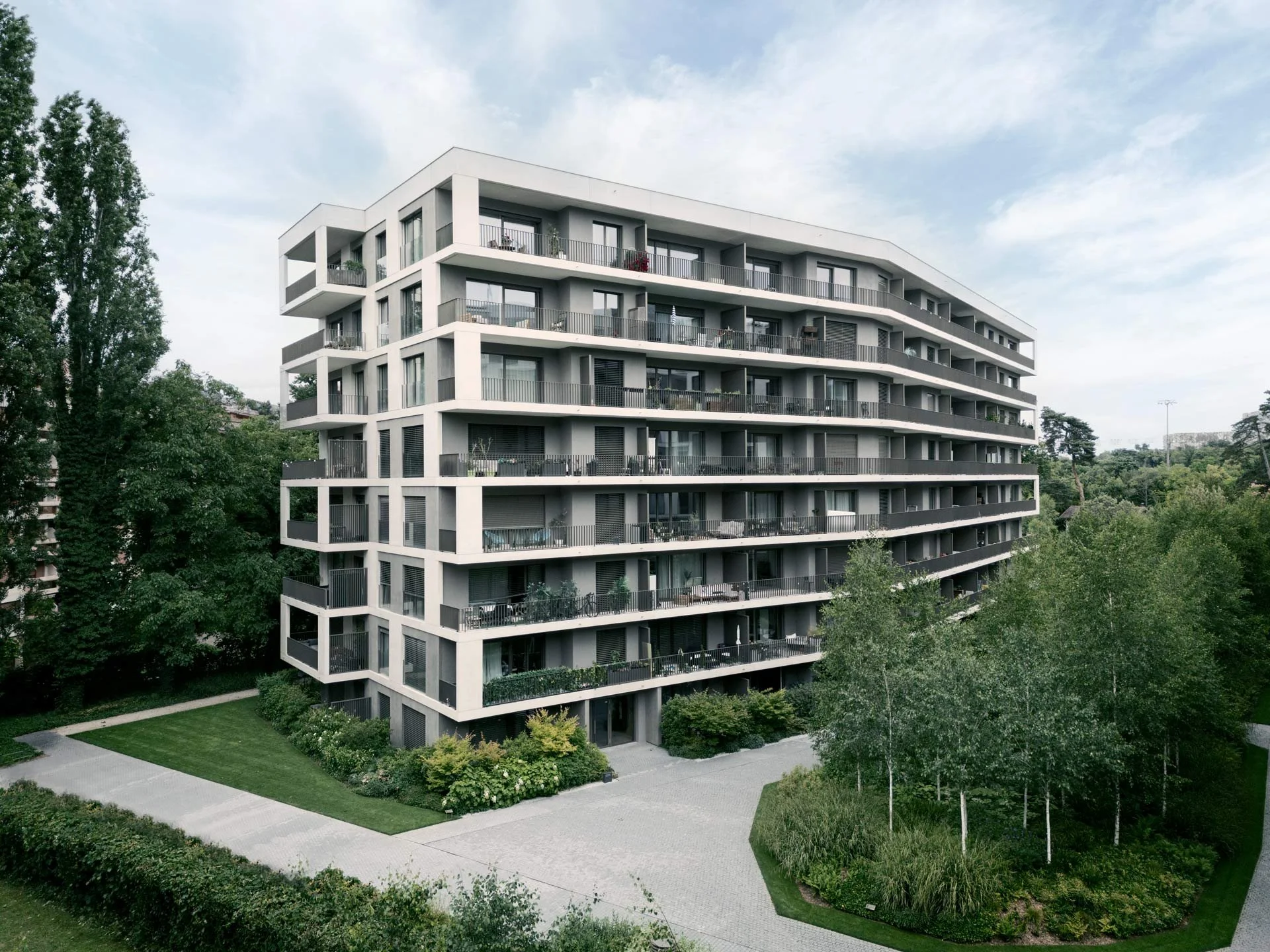 Immeuble résidentiel moderne avec plusieurs balcons et largement entouré d'arbres et de espaces verts.