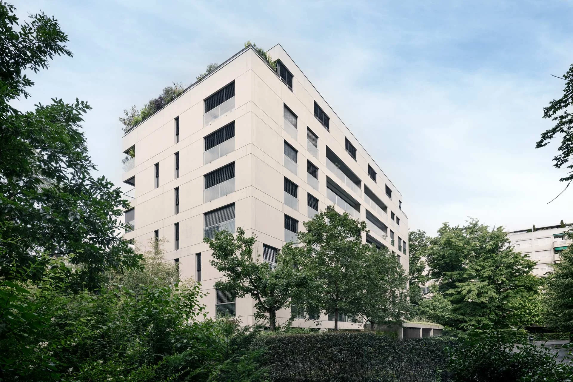 Un bâtiment moderne blanc avec plusieurs étages, entouré d'arbres verts, dans un environnement urbain calme.