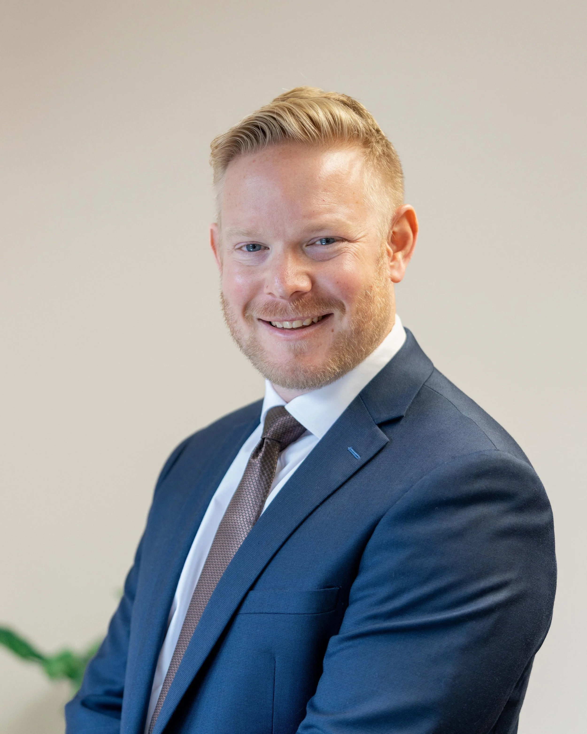 Smiling man in formal suit with a white shirt and brown tie, posing against a neutral background.