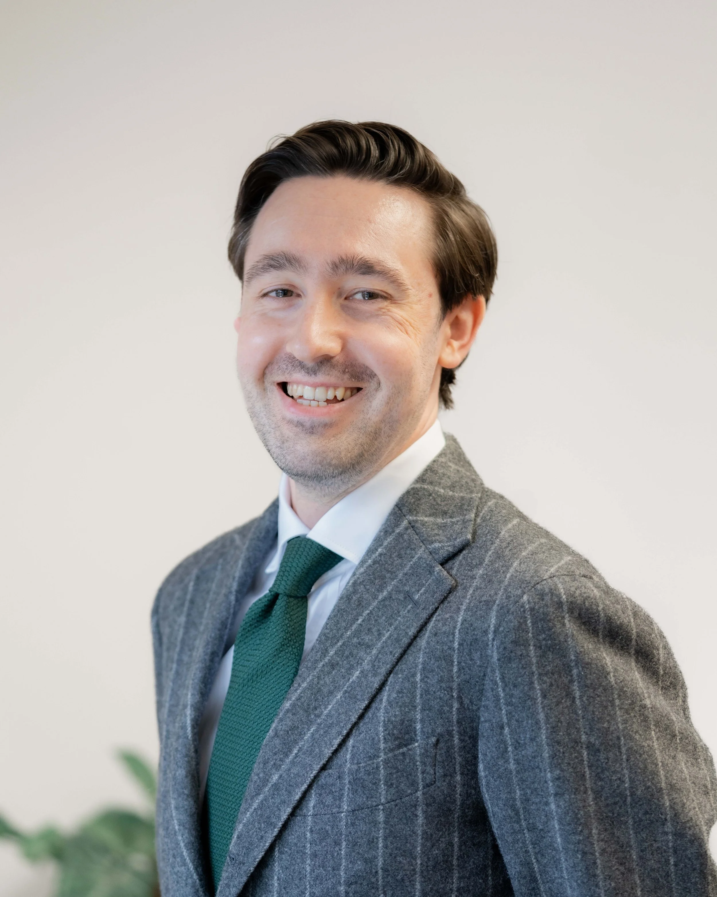 A man with dark hair wearing a gray pinstripe suit, white shirt, and green tie smiling at the camera.