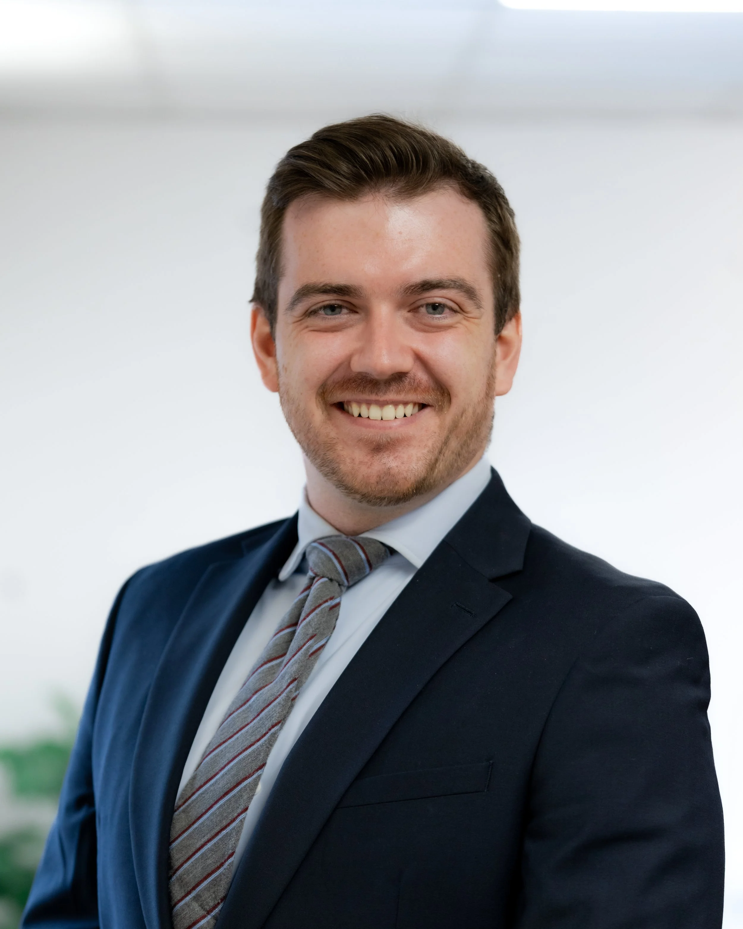 Headshot of a smiling young man with brown hair, wearing a dark suit, white shirt, and a gray striped tie, standing outdoors with a blurred background.